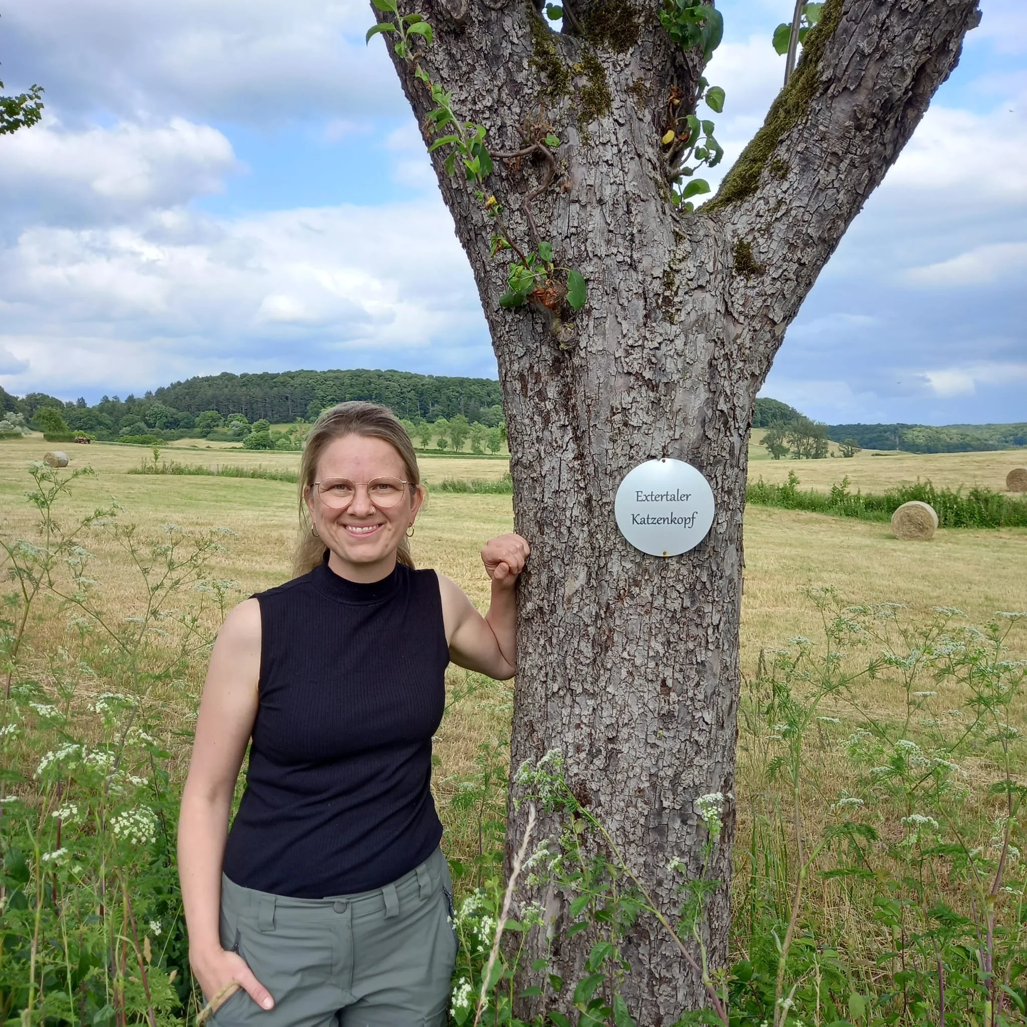 Eine lächelnde Frau steht neben einem Baum mit einer Tafel, die 'Extertaler Katzenkopf' heißt. Die Landschaft ist offen mit Feldern, Wiesen und einigen Baumgruppen im Hintergrund unter einem bewölkten Himmel.