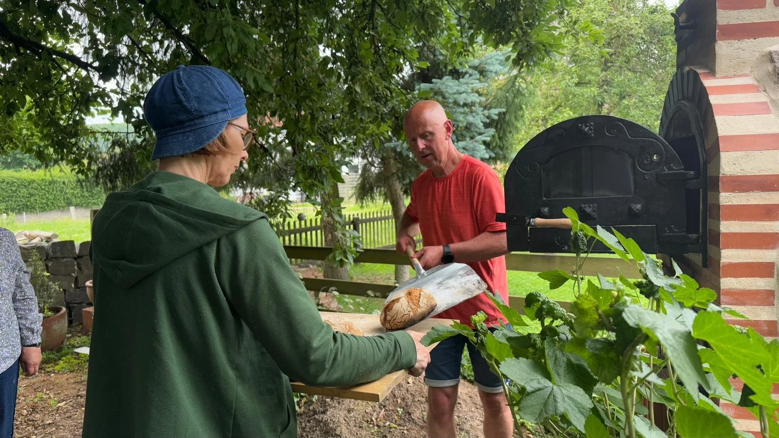 Zwei Menschen backen Brot im Garten, ein Mann schneidet Brot mit einem Messer, während eine Frau es auf einen Schneidebrett hält, um es zu servieren, bei sommerlichem Wetter unter einem Baum.