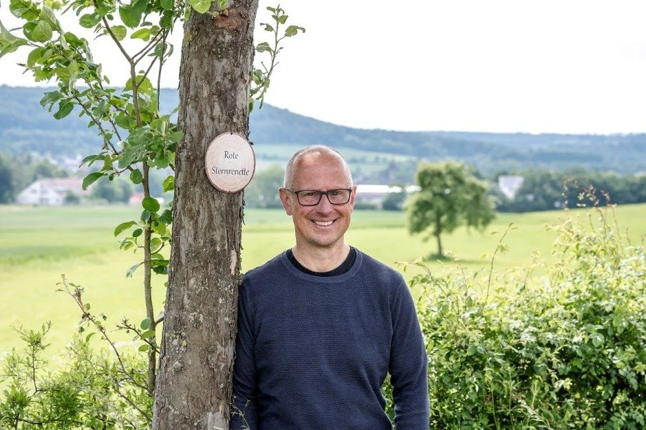 Ein lachender Mann mit Brille steht im Freien neben einem Baum in einem grünen, ländlichen Gebiet mit Wiesen und Bäumen im Hintergrund.