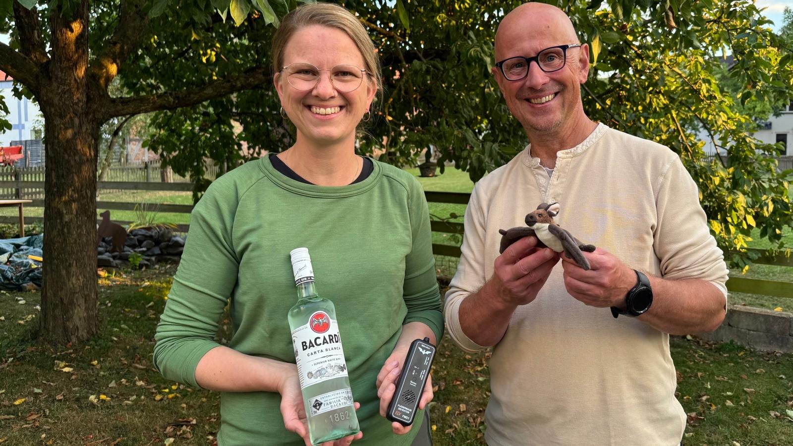 Zwei lachende Menschen, eine Frau mit Brille in grünem Sweatshirt und ein Mann mit Brille in hellem Pullover, halten eine Flasche Bacardi und eine Stofffigur in Form eines Affen vor einem Baum im Freien.