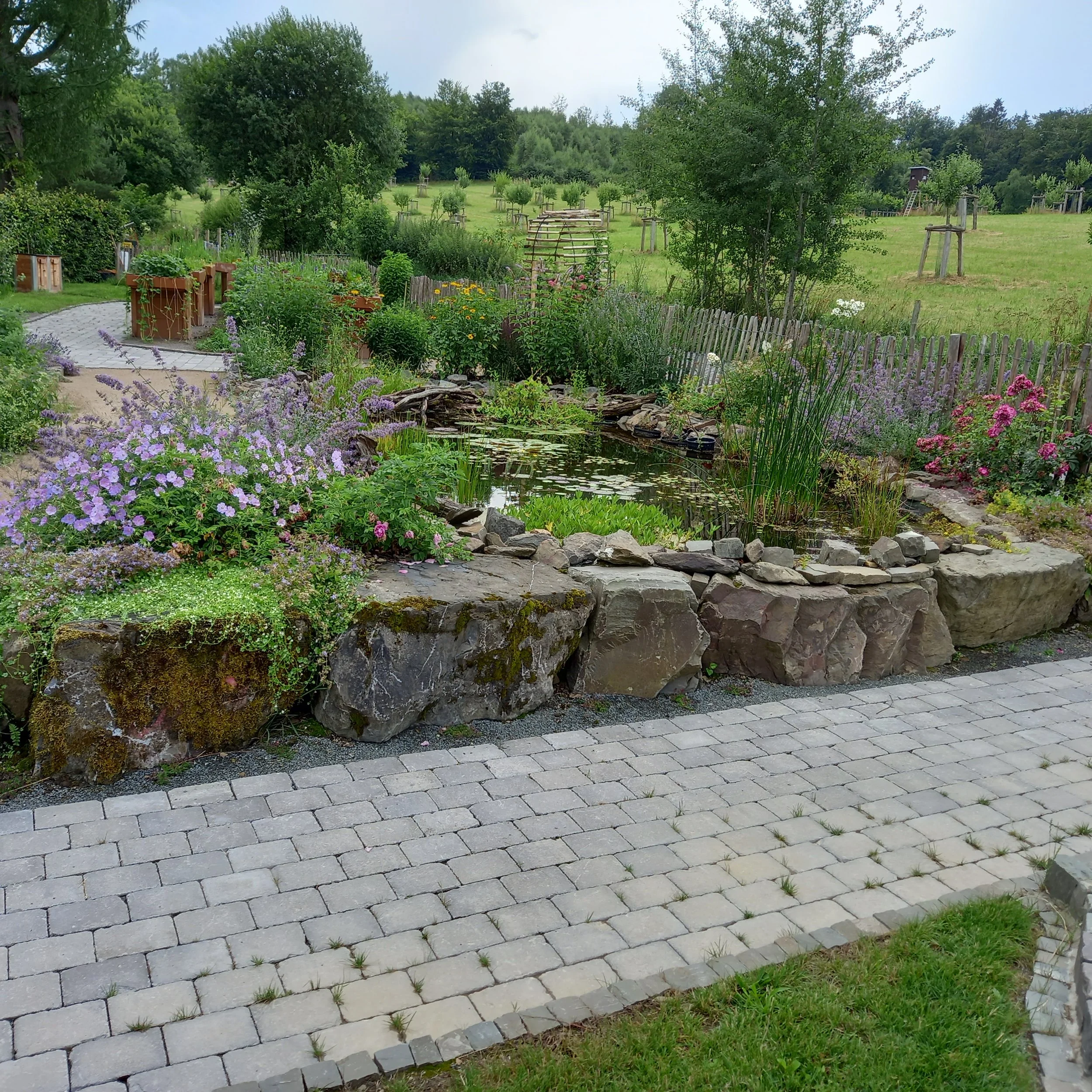 Garten mit Blumen, einem kleinen Teich, einem Kiesweg und Rasenfläche im Hintergrund auf einer bewölkten Wetter