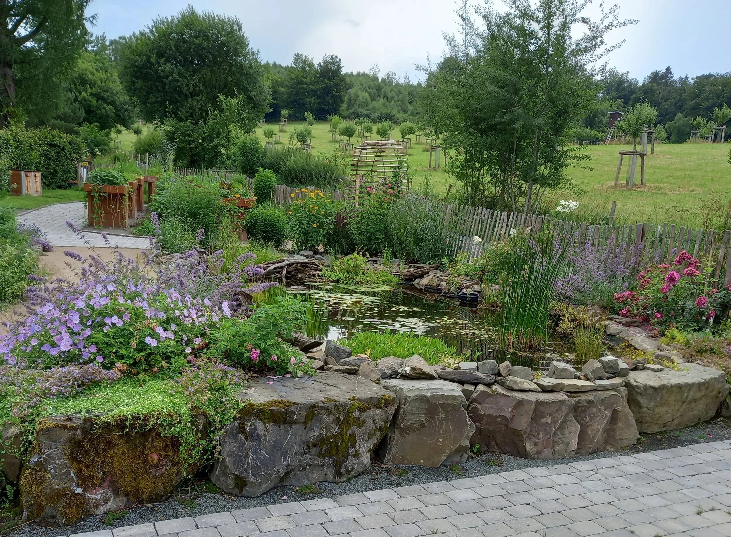 Ein gepflegter Garten mit Blumen, einem kleinen Teich und einem Kiesweg, umgeben von Bäumen und einem offenen Feld im Hintergrund.