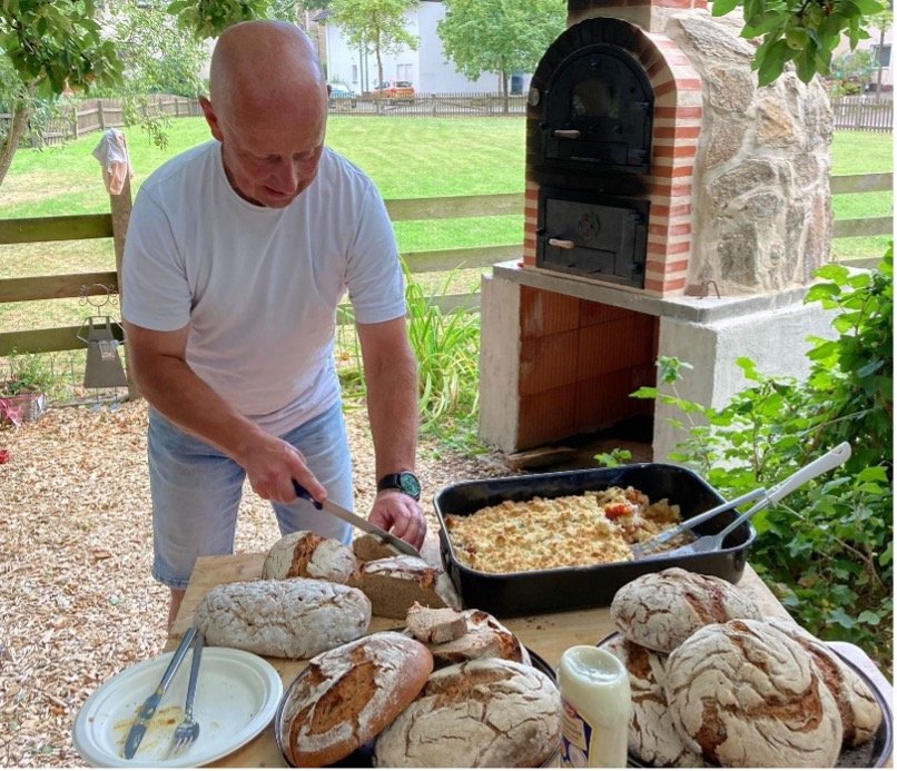 Ein älterer Mann schneidet Brot auf einer outdoor Frühstückstafel, mit mehreren Laib Brot, Aufstrich, Butter und einem Ofen im Hintergrund.