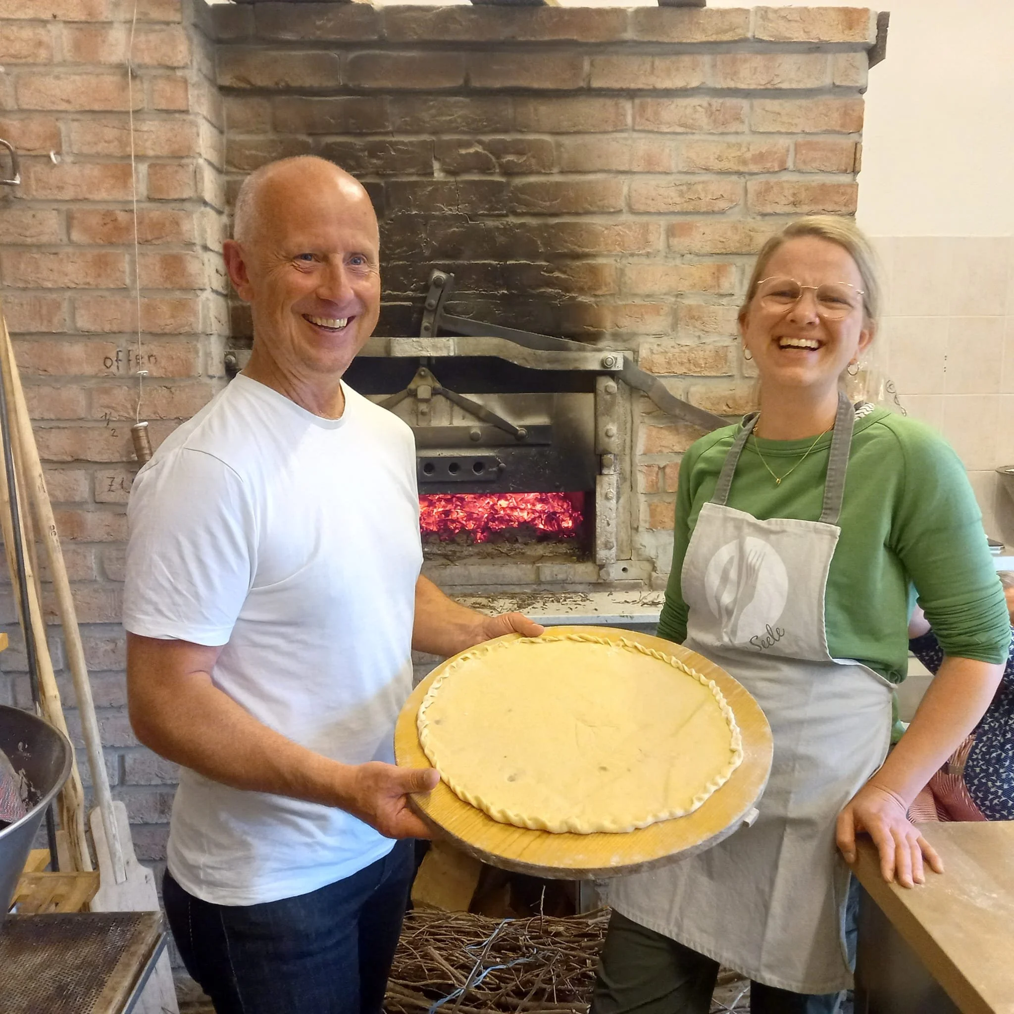 Zwei Menschen, ein Mann und eine Frau, stehen vor einem Backofen mit glühenden Kohlen und halten einen großen, ungebackenen Pizzateig auf einem Holzbrett. Beide lachen und schauen in die Kamera.