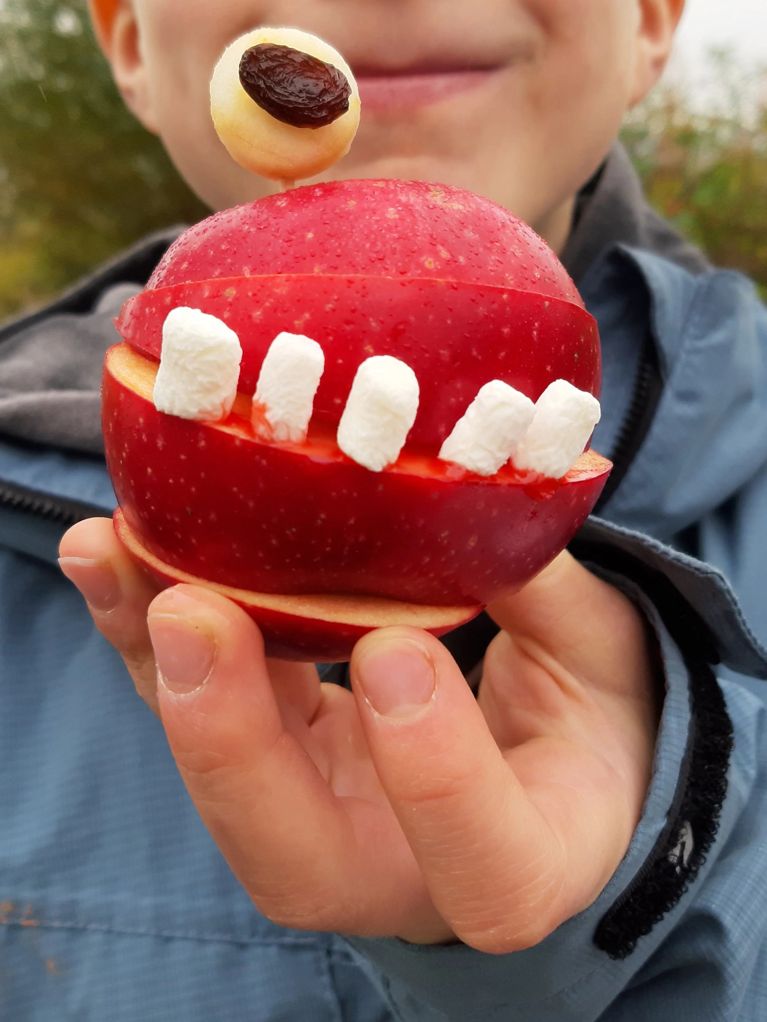 Ein Junge hält einen Apfel, der wie ein großer Mund gestaltet ist, mit Marshmallows als Zähne, und eine kleine Pizza mit einer Olive als Augenpartie darauf.