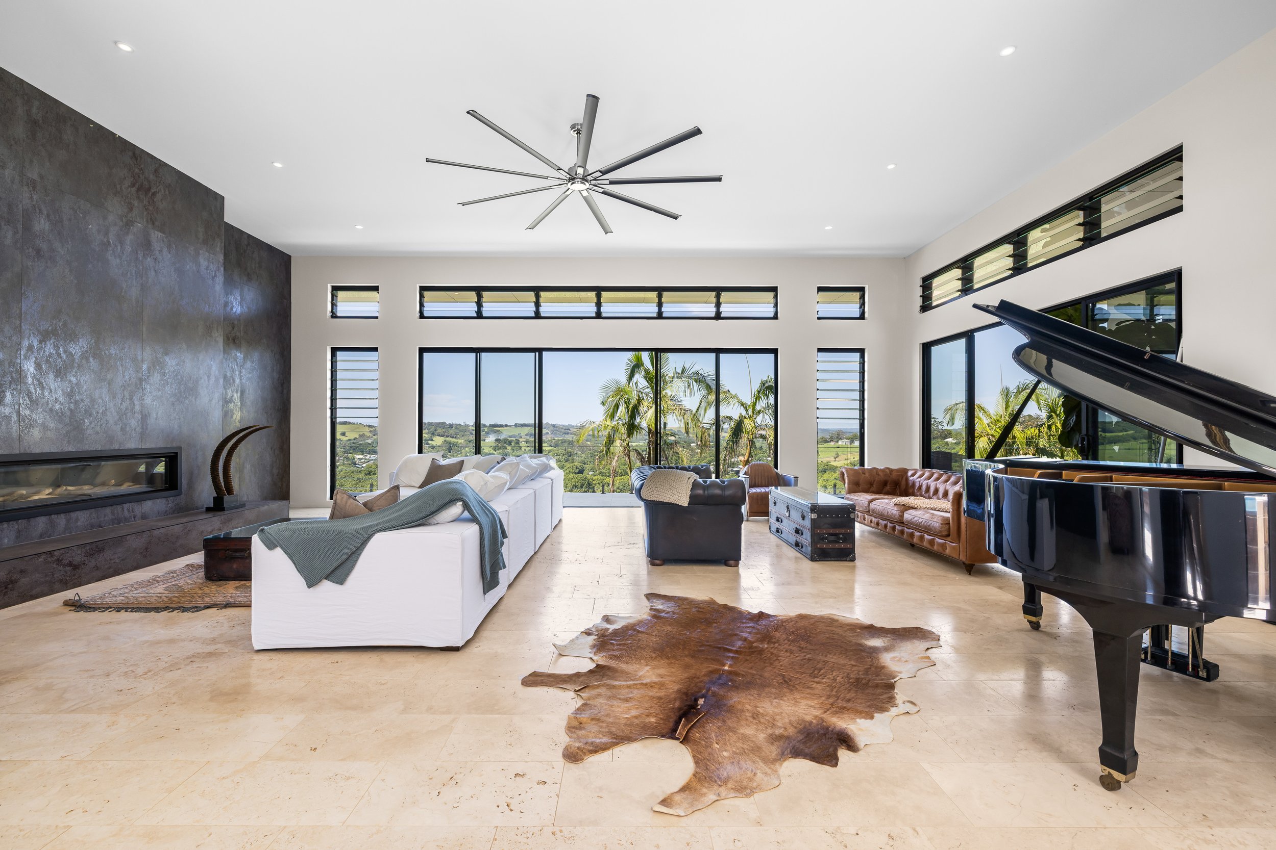 Living room with large windows, sectional sofa, armchair, cowhide rug, grand piano, and scenic view of trees and landscape.