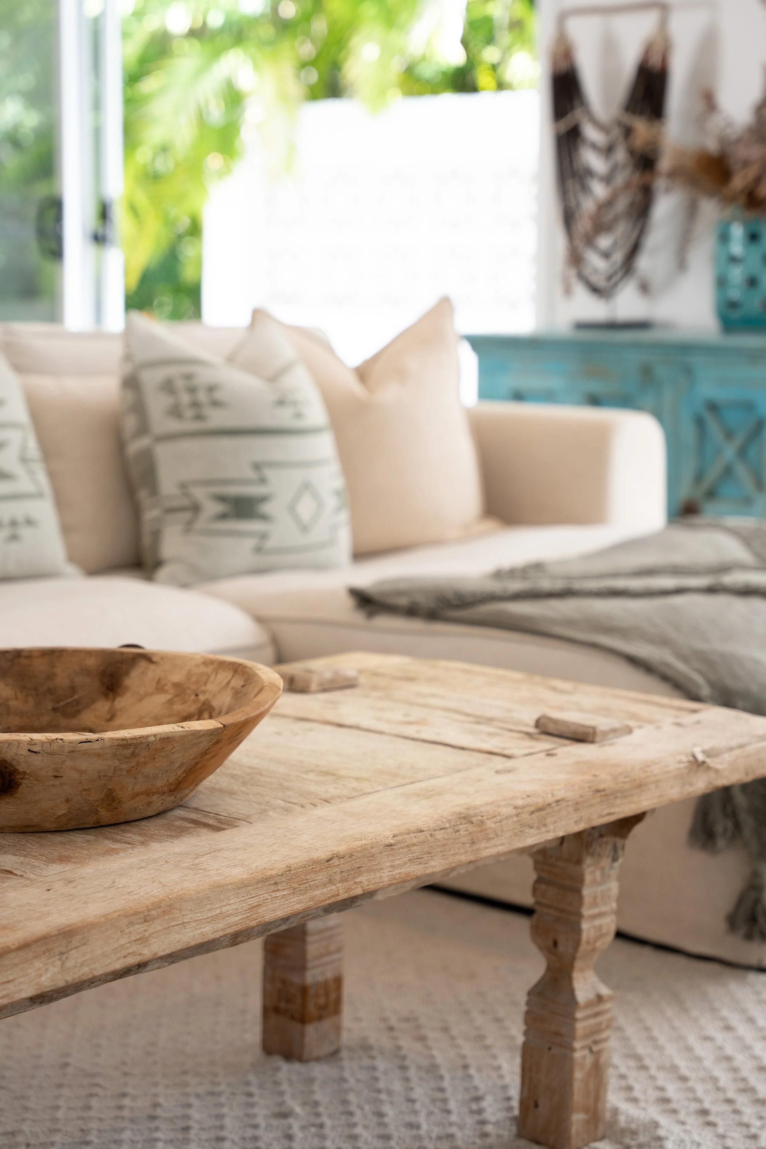 Close-up of a rustic wooden coffee table in a cozy living room with a white sofa and patterned pillows, a textured throw blanket, and decorative wall art in the background.