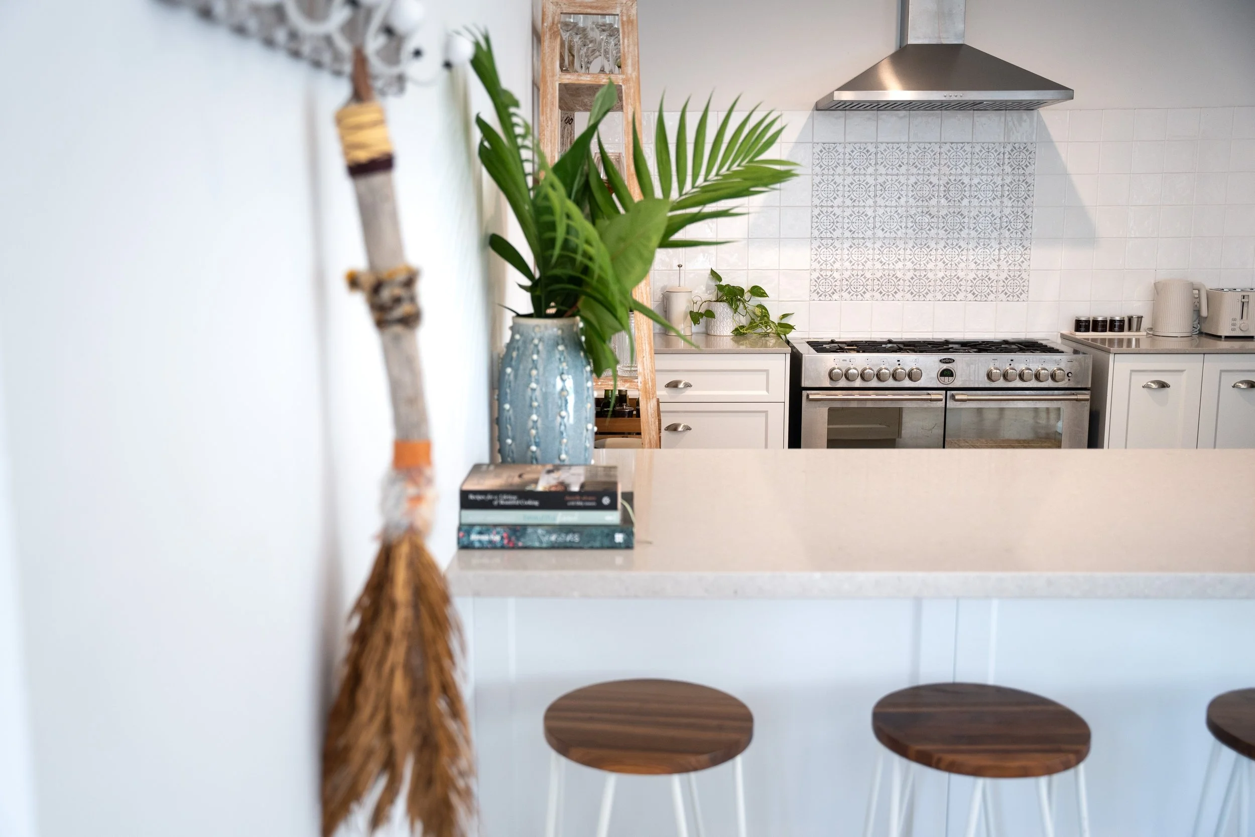 A kitchen with a white island counter, a large green plant in a blue vase on the counter, some books, and wooden bar stools. In the background, there is a stove with a stainless steel range hood, a white tiled backsplash with a decorative pattern, and white cabinets with a kettle, toaster, and spice jars.