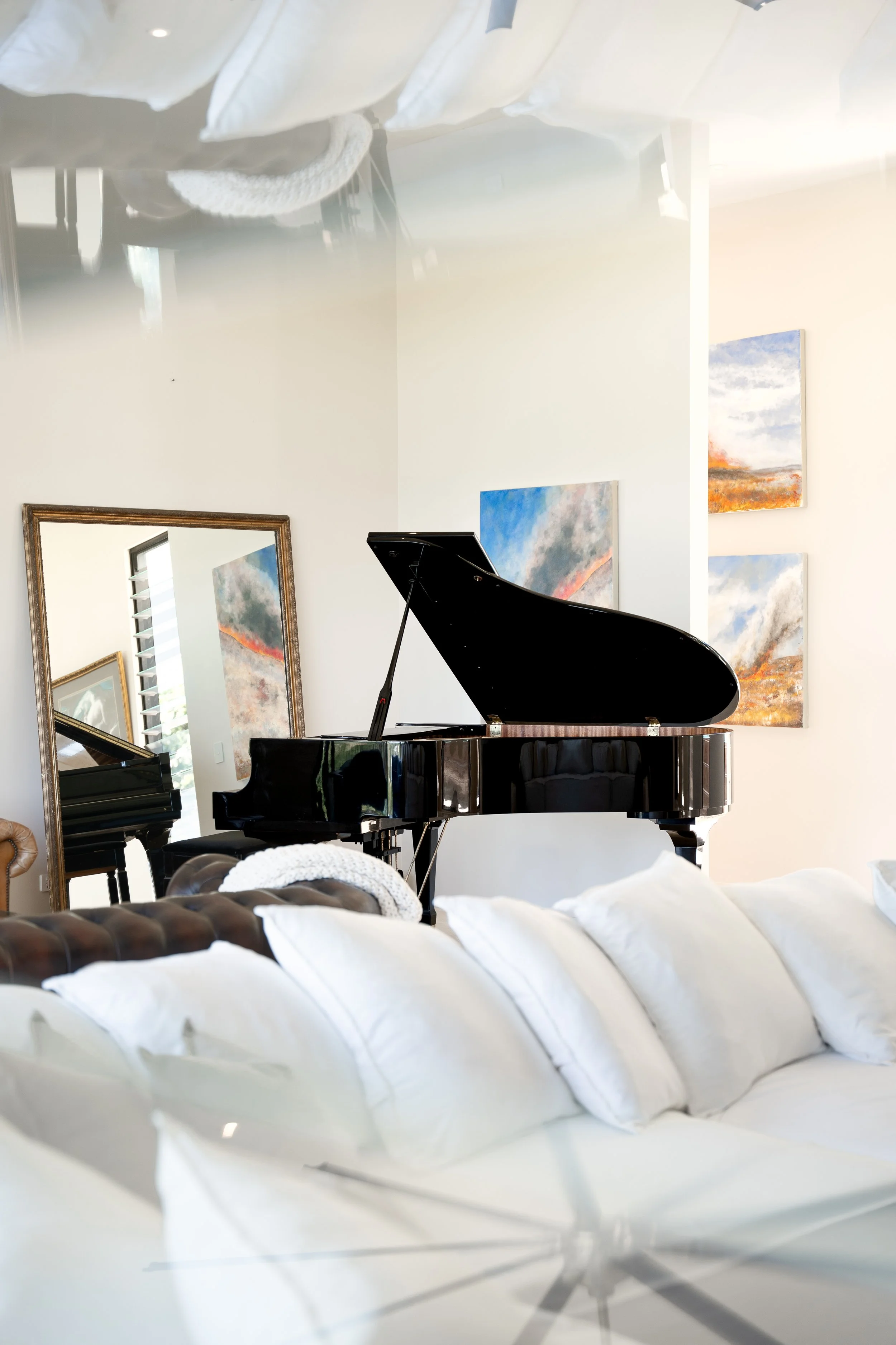 A black grand piano in a modern living room with white walls, a large mirror, and framed abstract landscape paintings.