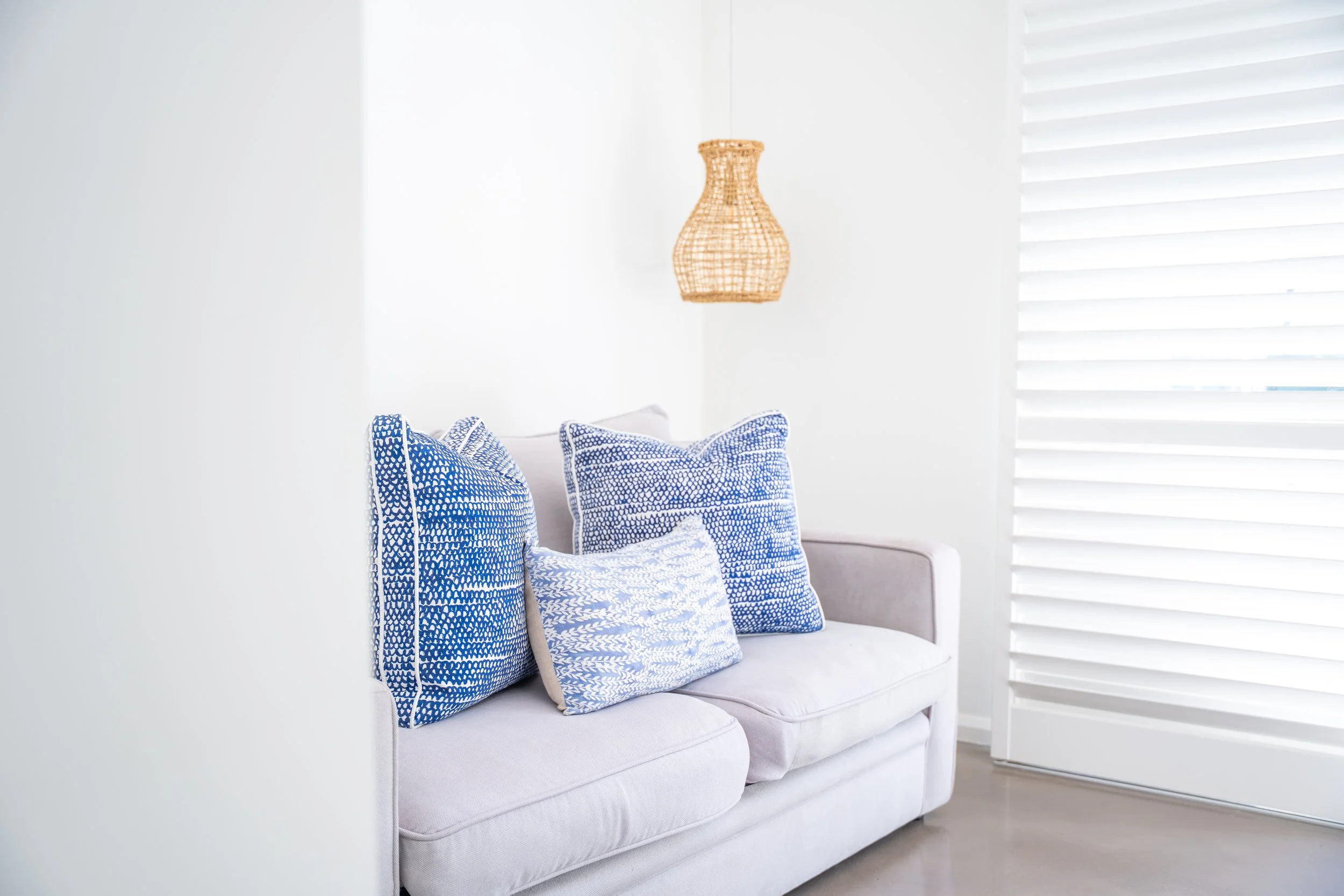A white sofa with three blue patterned pillows, a wicker wall lamp, and white plantation shutters partially covering a window.