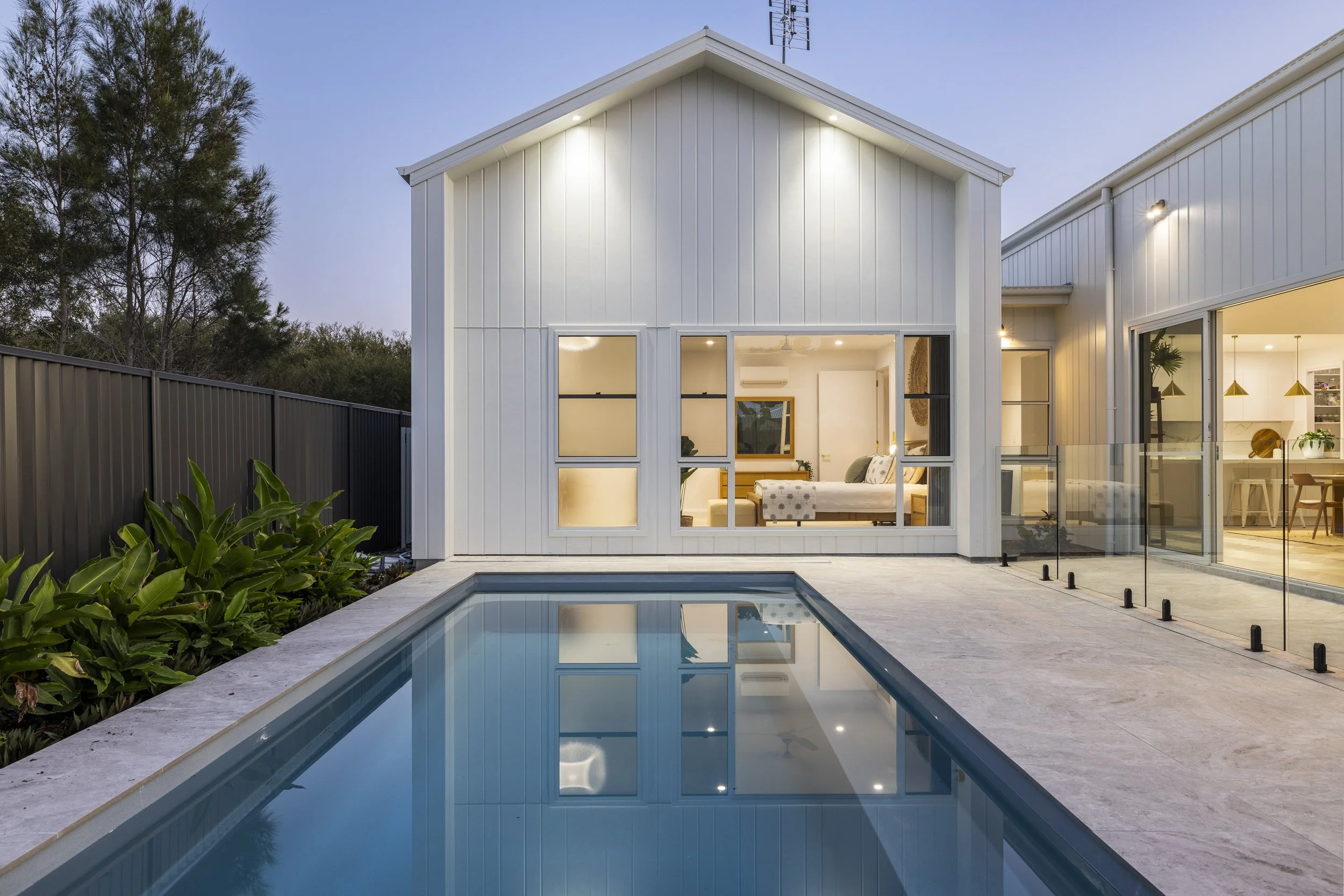 Modern white house with large windows, outdoor pool, glass fencing, and a patio area, illuminated during dusk.