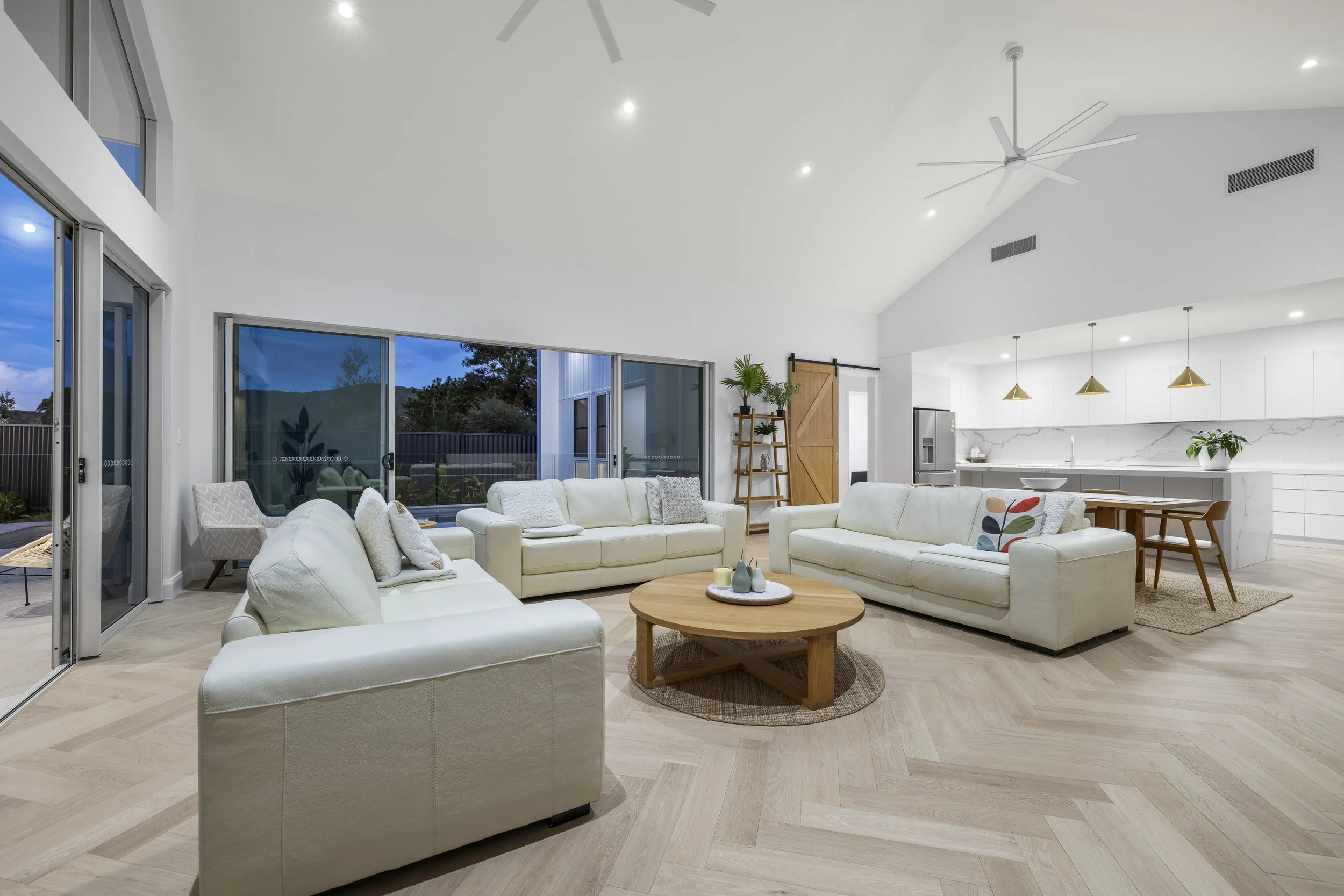A spacious, modern living room with white sofas, a round wooden coffee table, a large window with sliding doors, and an open kitchen area with white cabinets and a marble backsplash.