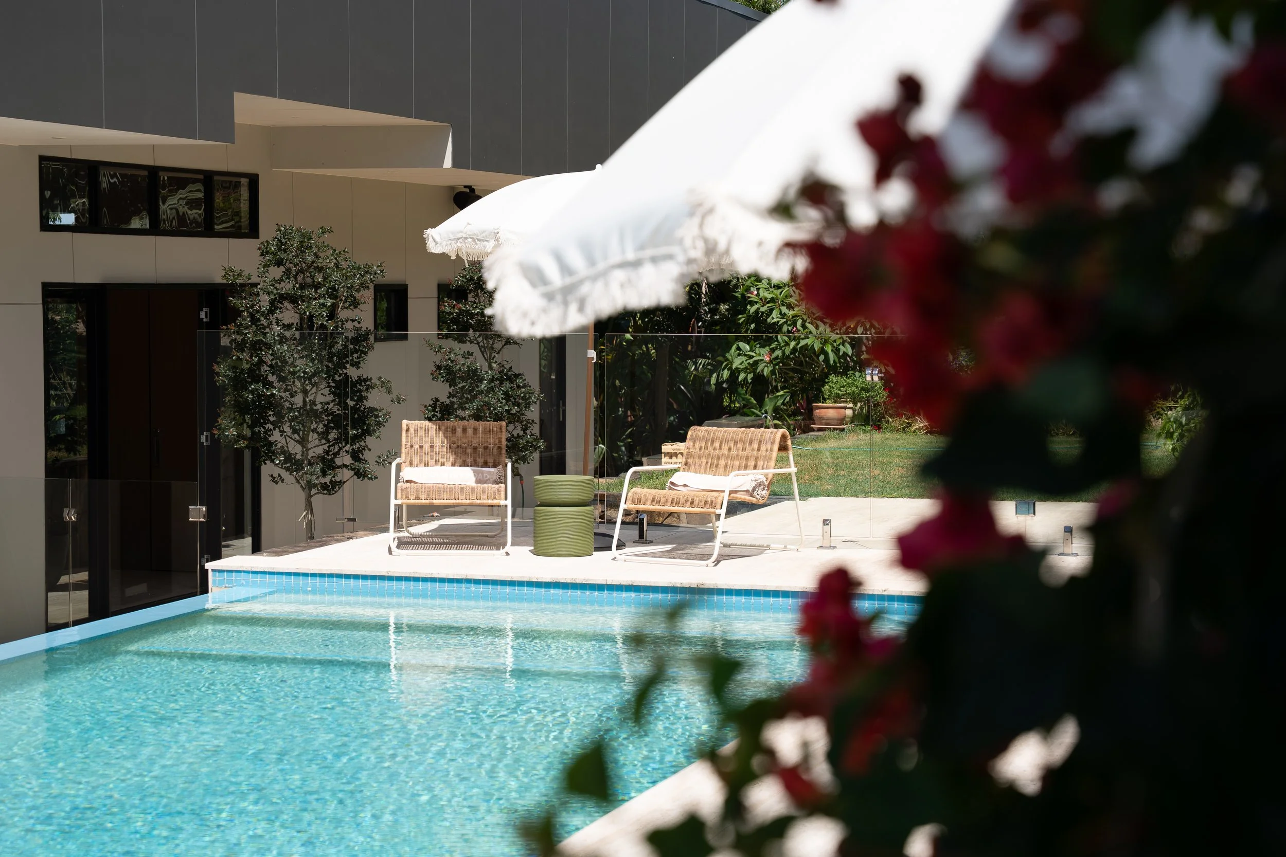 A backyard swimming pool with a patio area featuring two wicker chairs, a white umbrella, and a small green side table. There are trees and greenery in the background, and pink flowers partially obstruct the view in the foreground.
