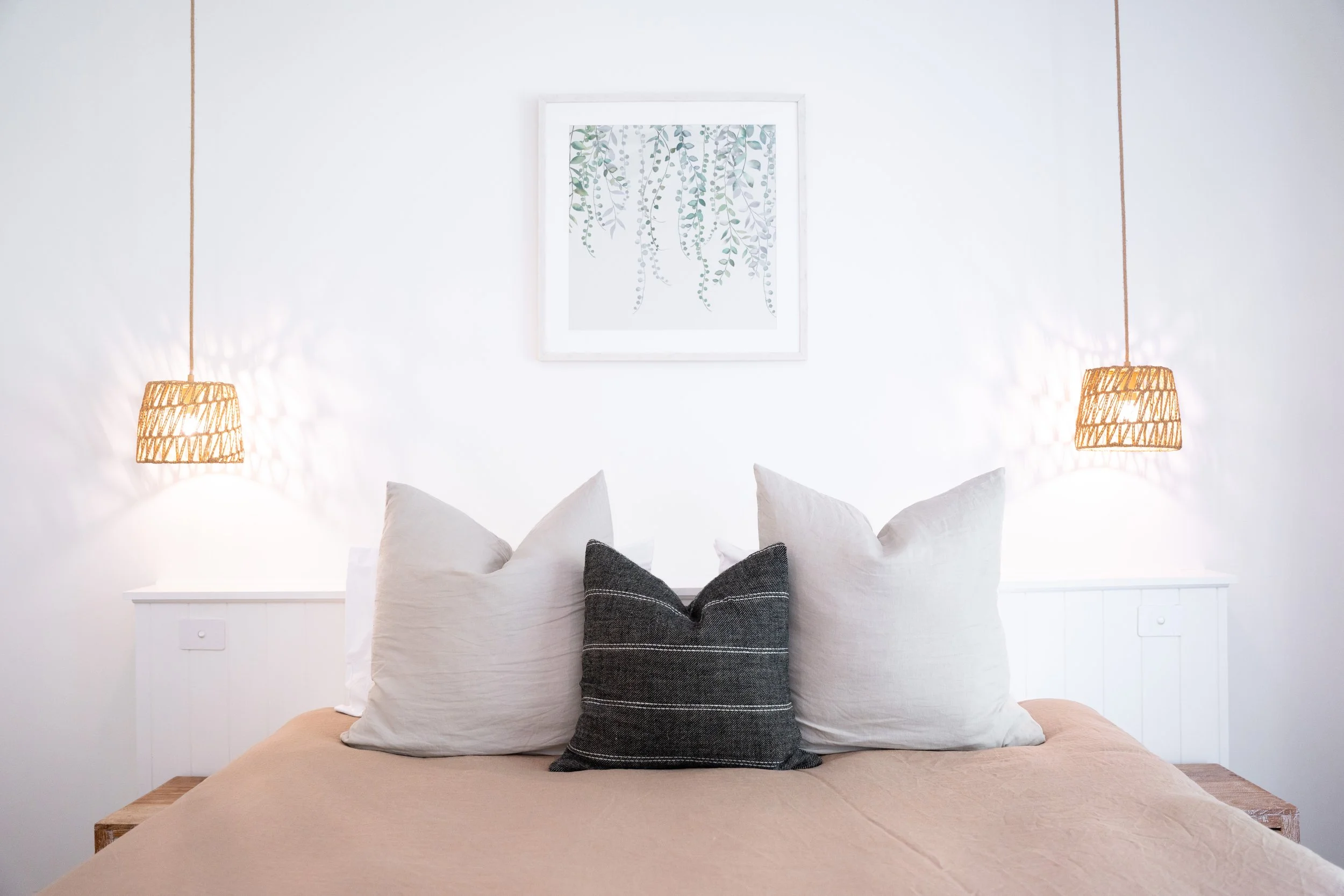 A neatly made bed with beige bedding, four large pillows including one black with white stripes, framed botanical artwork on white wall, two hanging wicker lamps on either side.