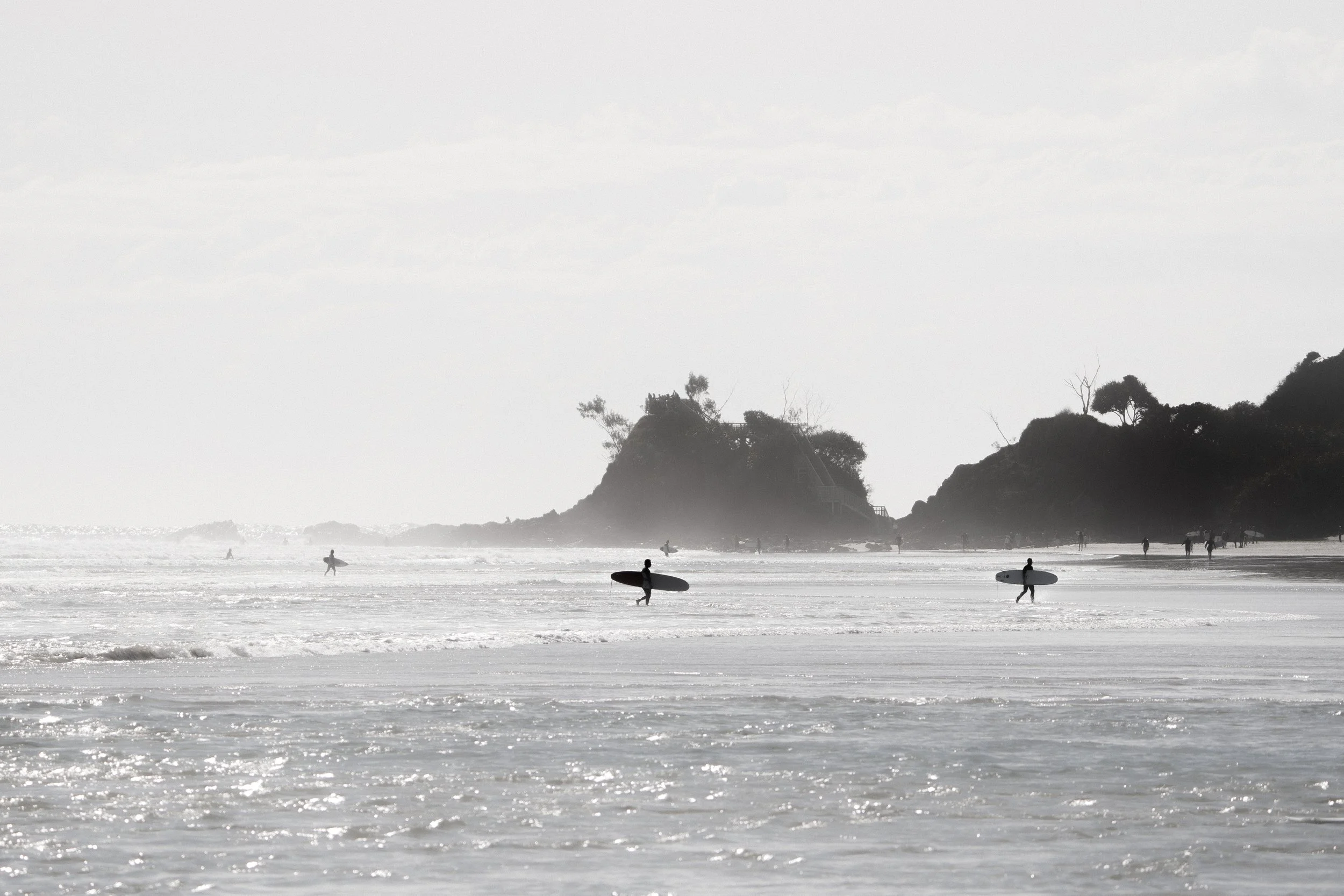 People surfing and walking on the beach near rocky hills under cloudy skies