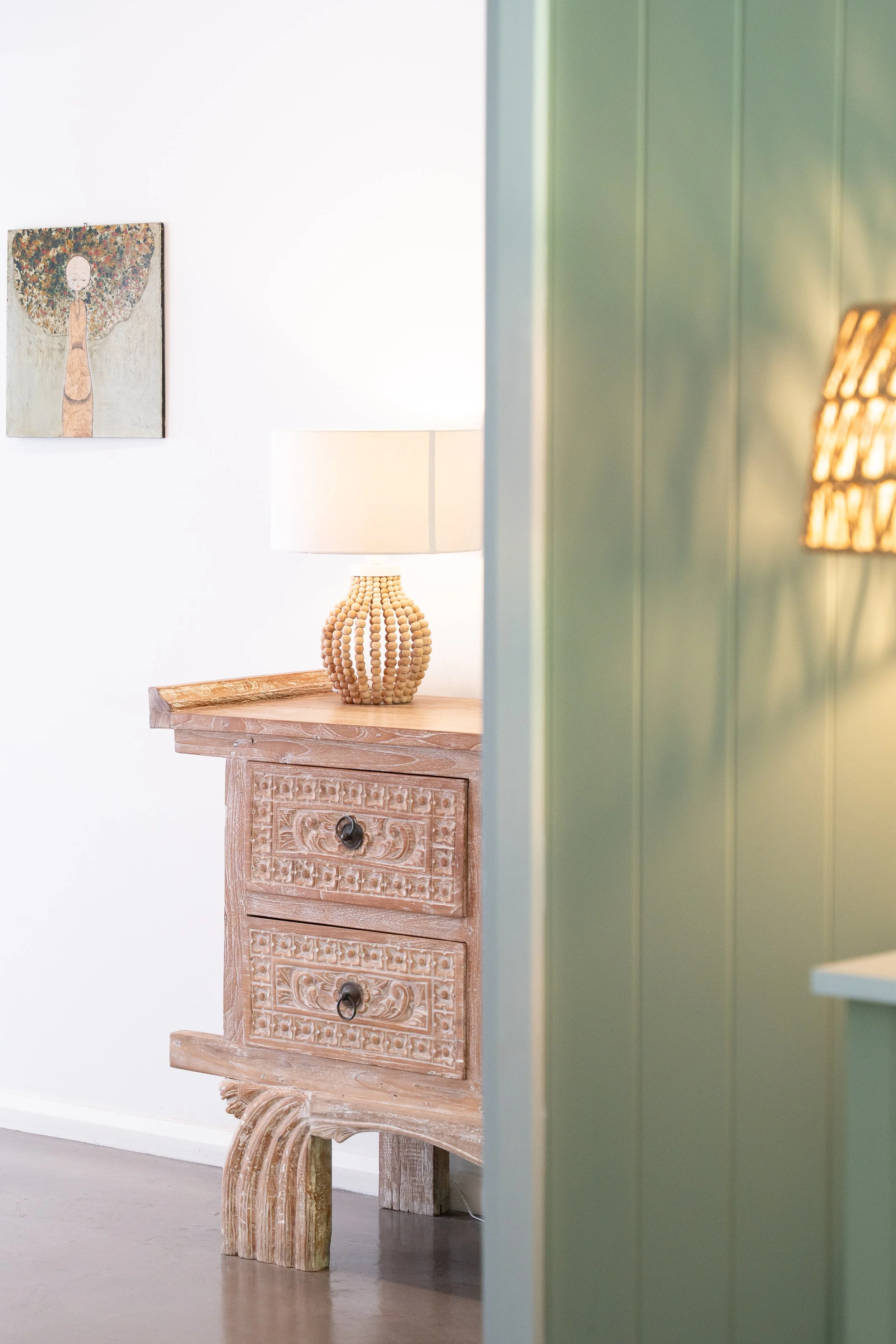 A wooden carved dresser with two drawers, a beige table lamp, a green wall, and a painting on a white wall. Part of a yellow lamp is also visible.