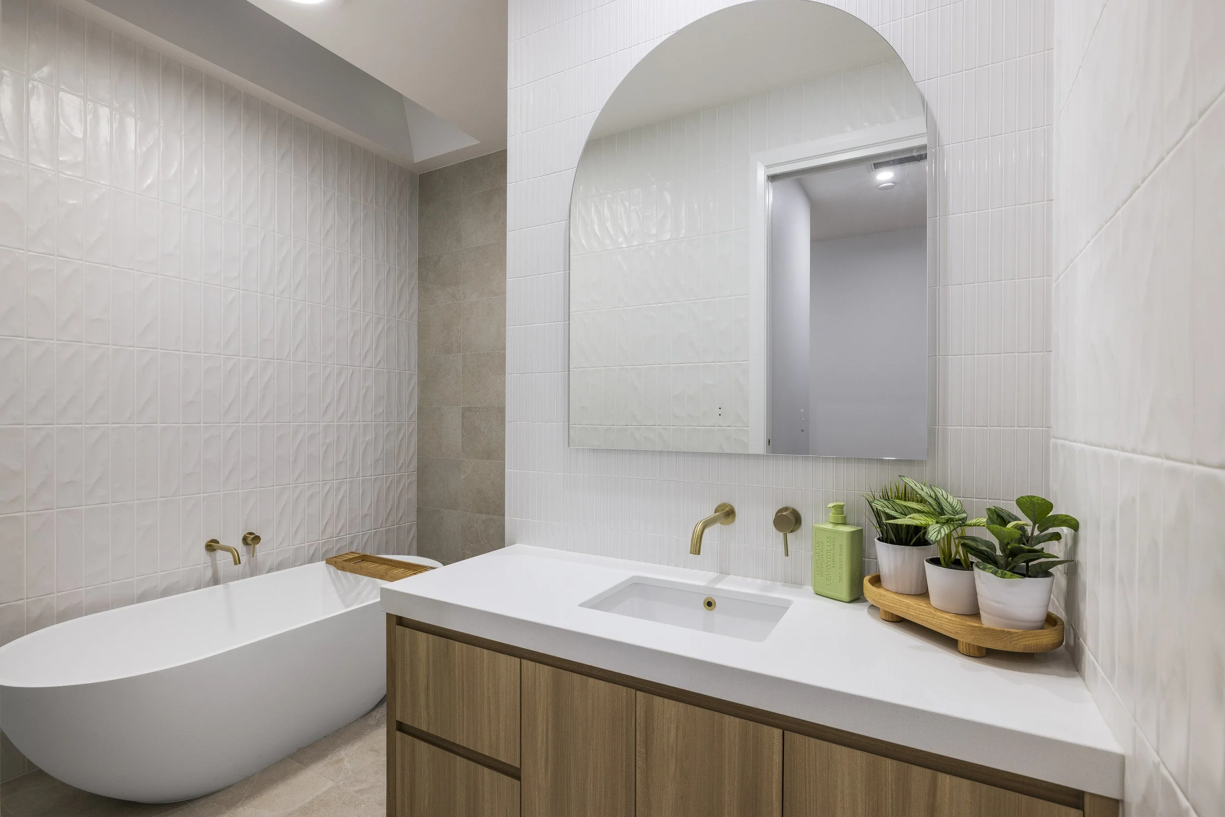 Modern bathroom with freestanding bathtub, wooden vanity with white countertop, large mirror, and several potted plants.