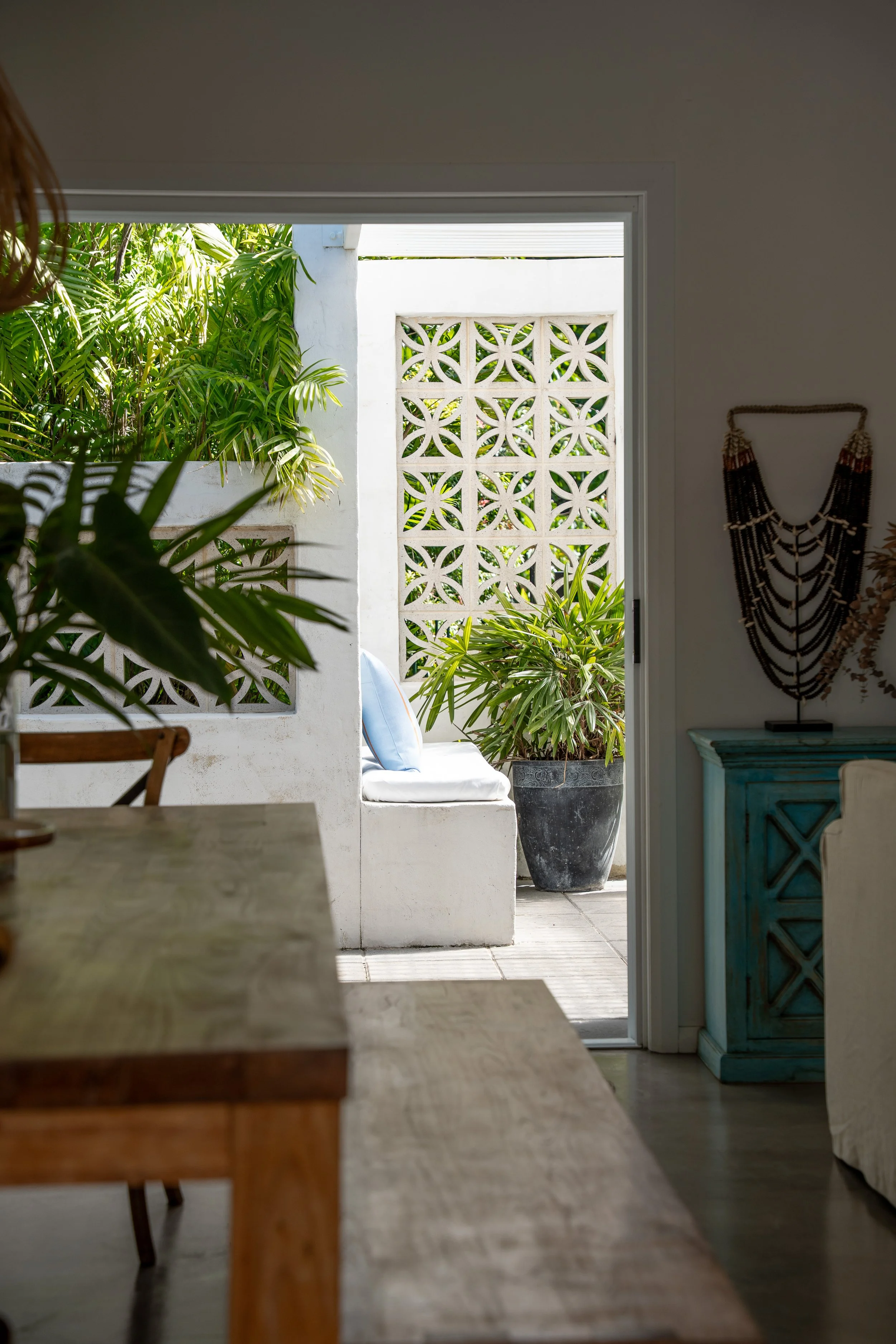 Indoor view looking out to a patio with potted plants, a white built-in bench with a blue pillow, and a decorative wall block with circular cutouts, featuring lush greenery and sunlight.
