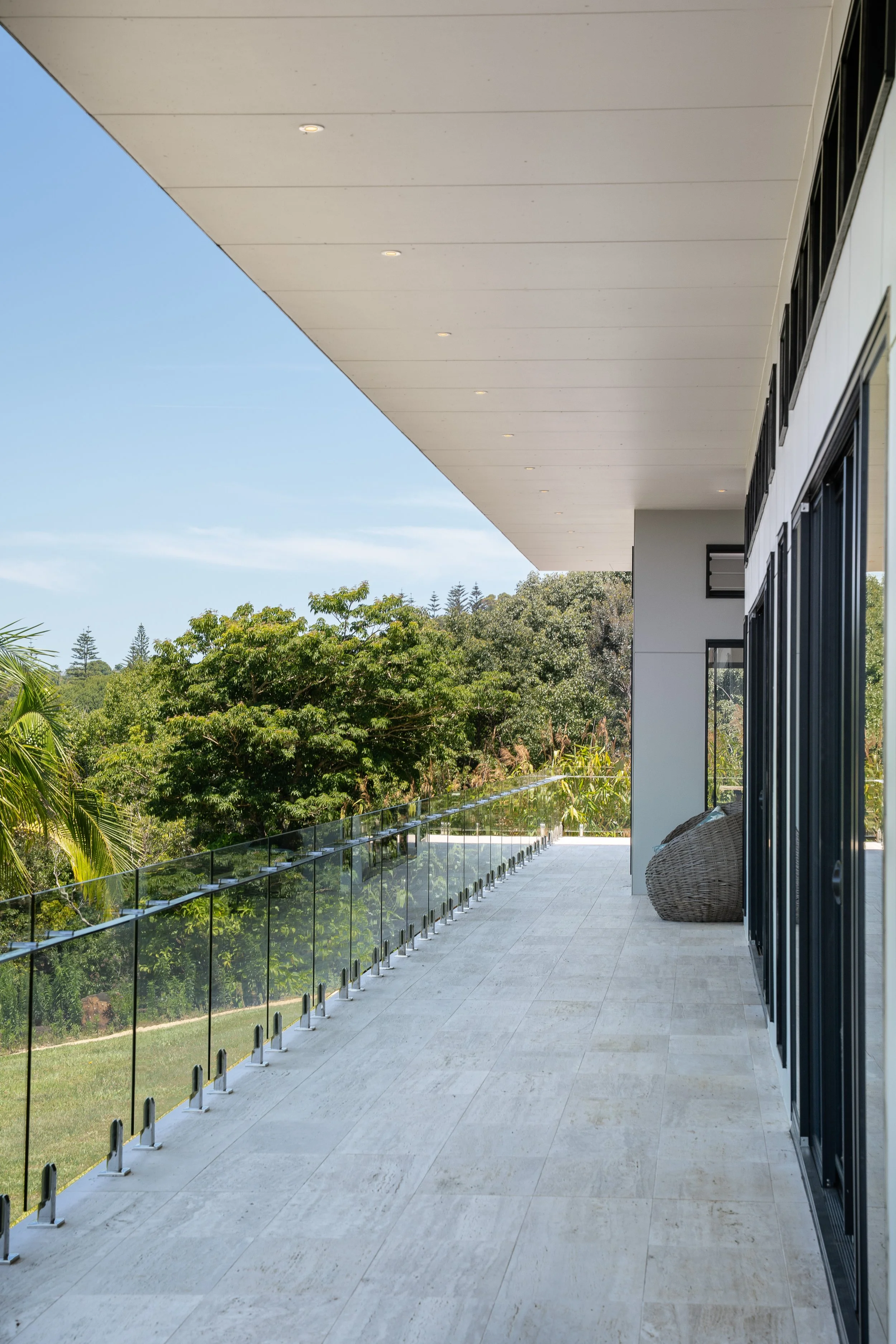 A modern balcony with glass railing overlooking lush green trees, with a wicker chair and sliding glass doors on the right.
