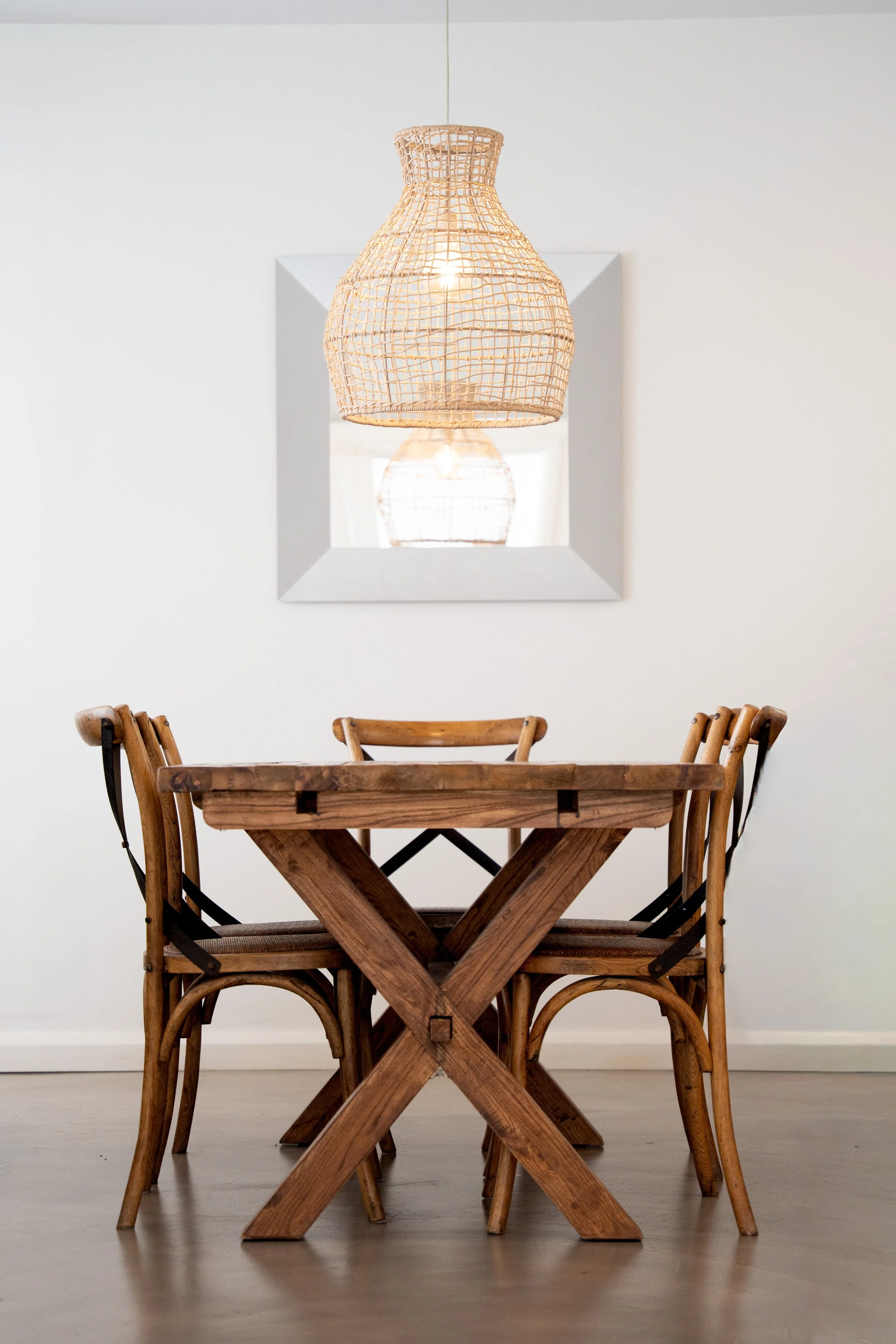 A wooden dining table with four matching chairs, a wicker pendant light hanging above, and a square mirror on the plain white wall reflecting the light fixture.