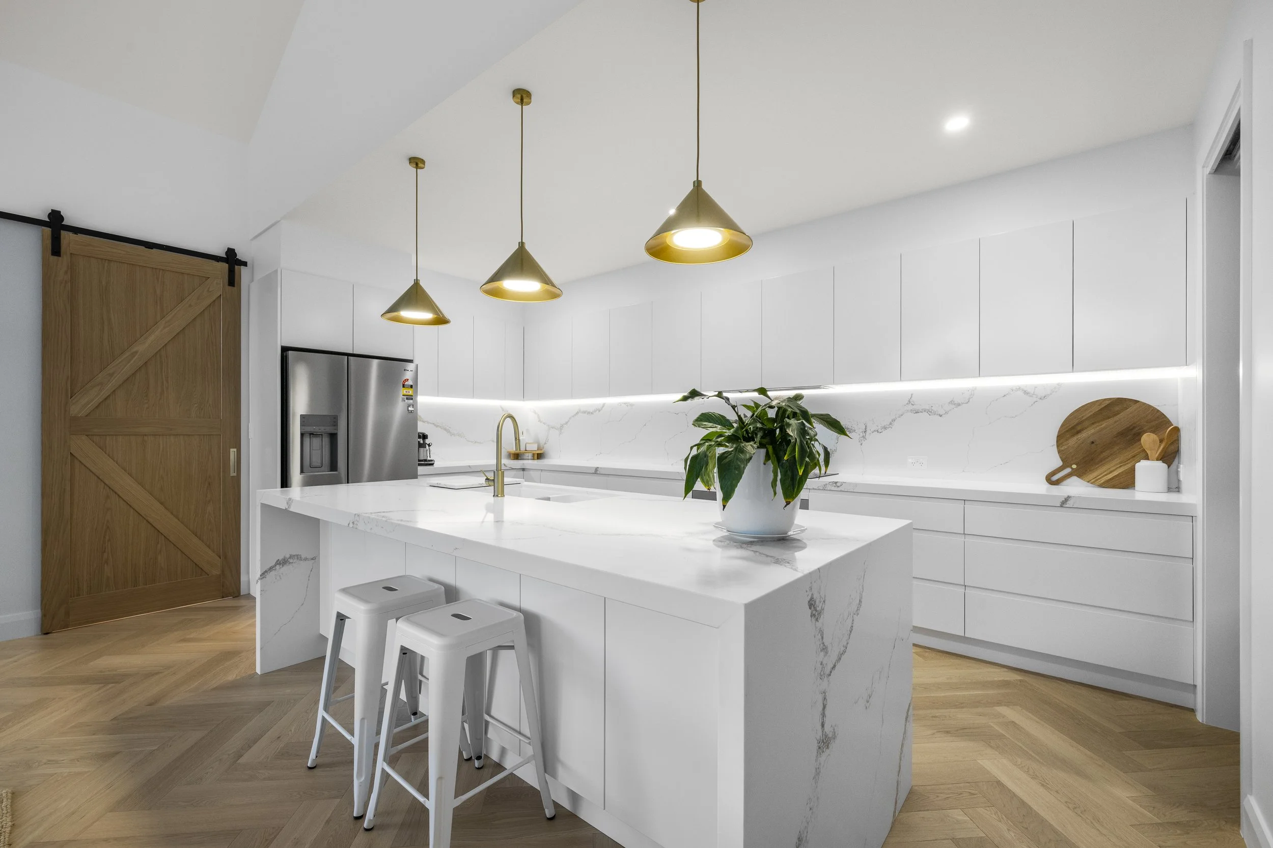 Modern white kitchen with marble countertops and backsplash, gold pendant lighting, a built-in refrigerator, a potted plant on the island, and wooden accents including a sliding barn door and cutting boards.