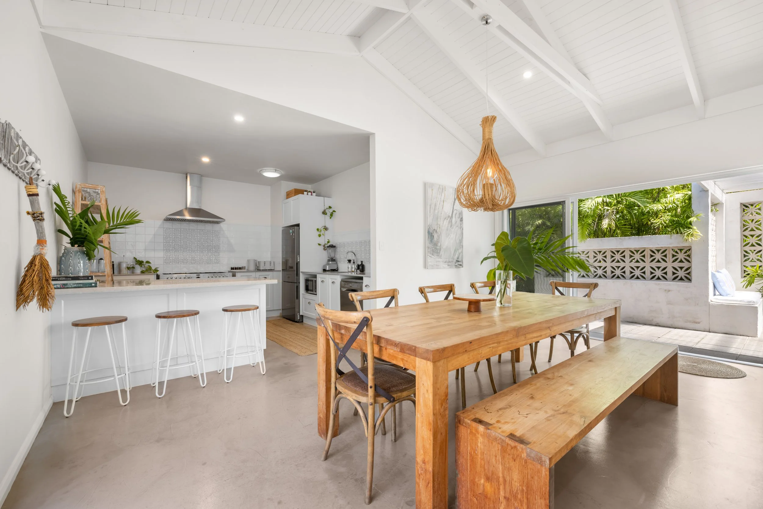 Open-concept kitchen and dining area with white walls, wooden dining table, benches, and chairs, large window with greenery outside, hanging wicker light fixture, potted plants, and minimal decor.