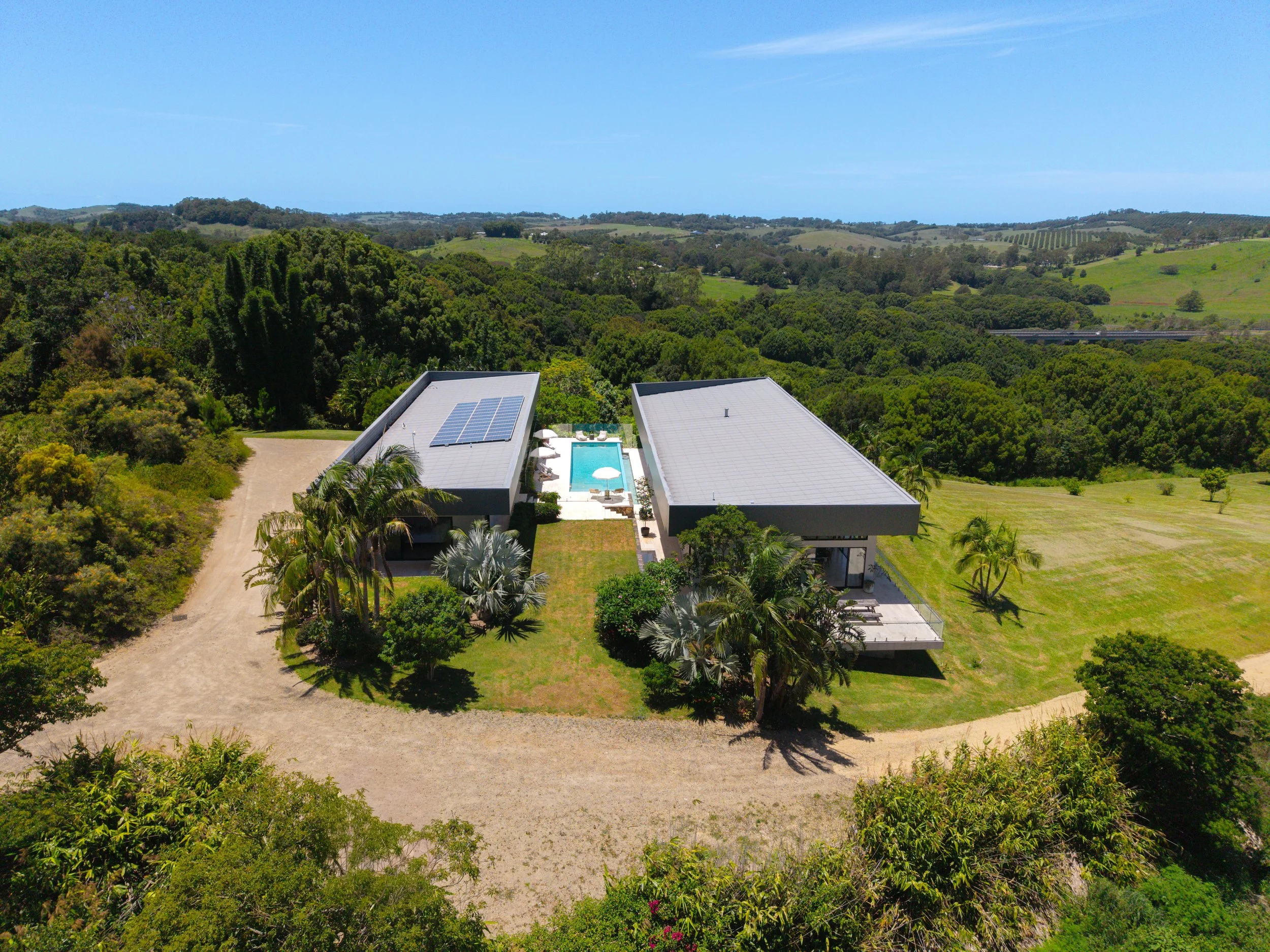 Aerial view of a modern house with solar panels, a swimming pool, and lush greenery in a rural area with rolling hills and trees.