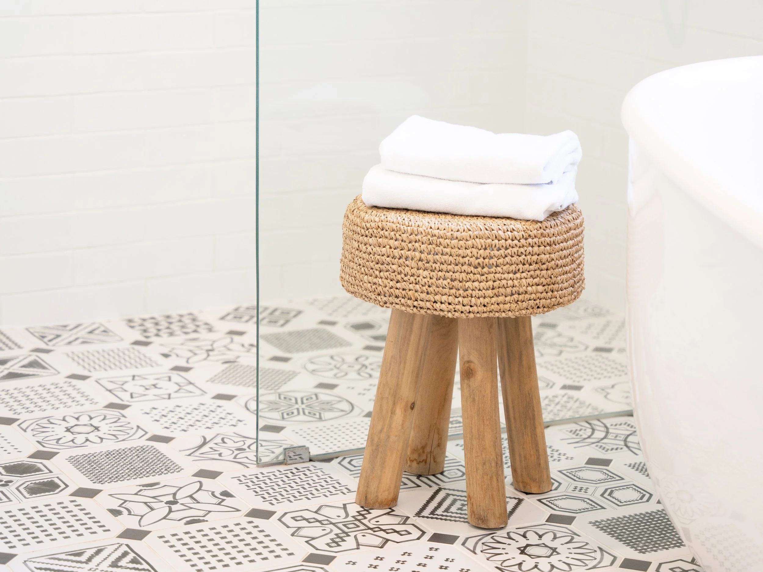 Bathroom with white tiled walls, black and white decorative tiled floor, a wooden stool with a woven top and folded white towels on it, and partial view of a white bathtub.