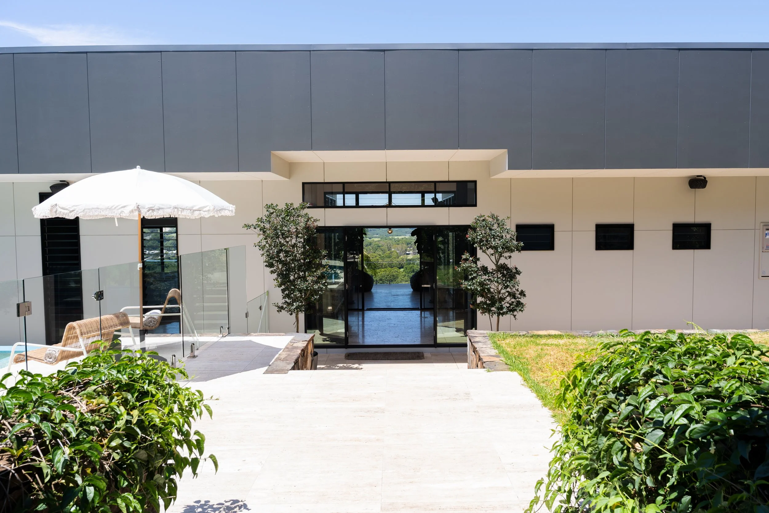 Modern building entrance with glass doors, two small trees, outdoor seating area with an umbrella, a grassy area, and bushes in the foreground.