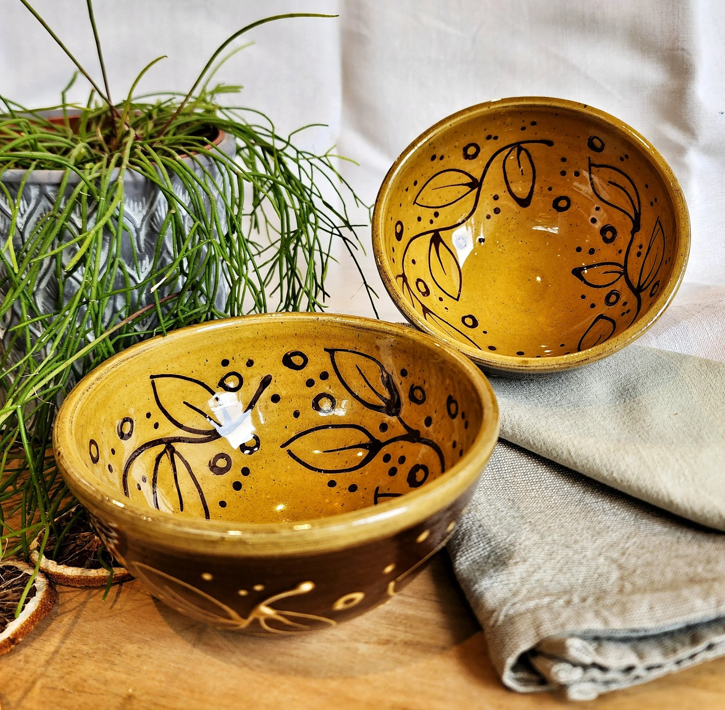 Two yellow ceramic bowls with black leaf and dot patterns, placed on a wooden surface with a green and beige cloth, and a potted green plant in the background.