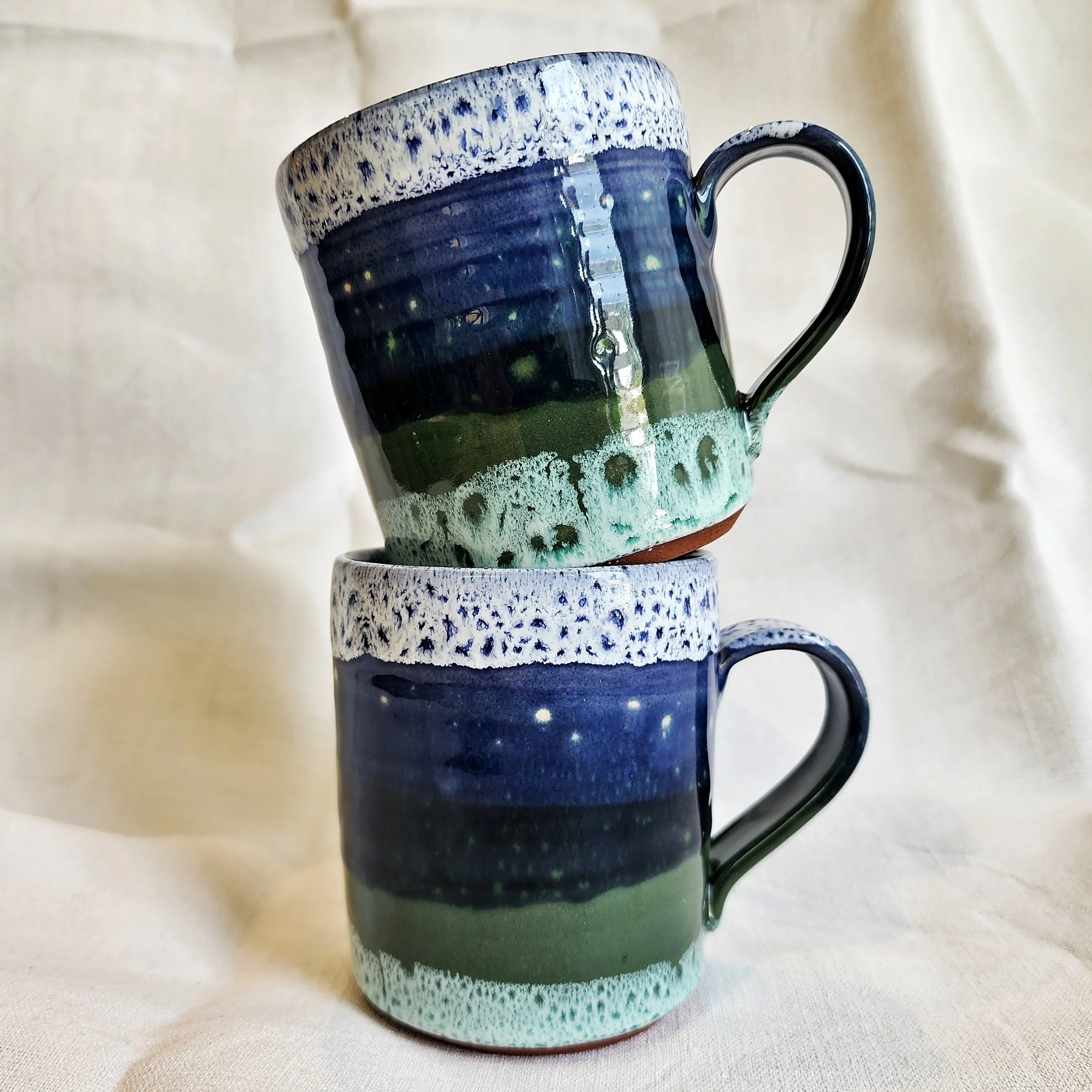 Two stacked ceramic mugs with galaxy-like and nature-inspired designs on a neutral fabric background.