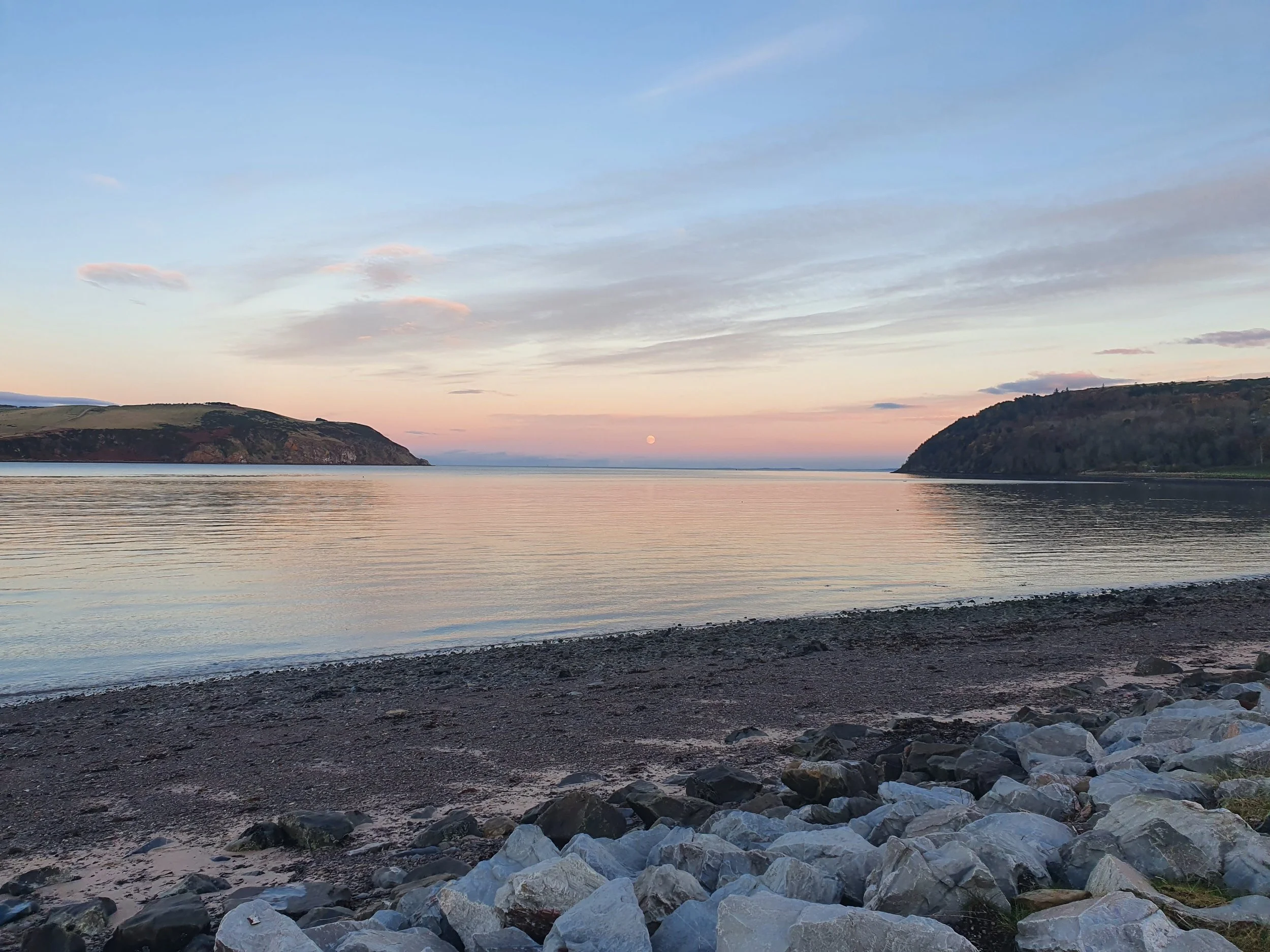 A serene coastal scene in Cromarty during sunset with a calm body of water, rocky beach in the foreground, and hills on either side under a pastel-colored sky.