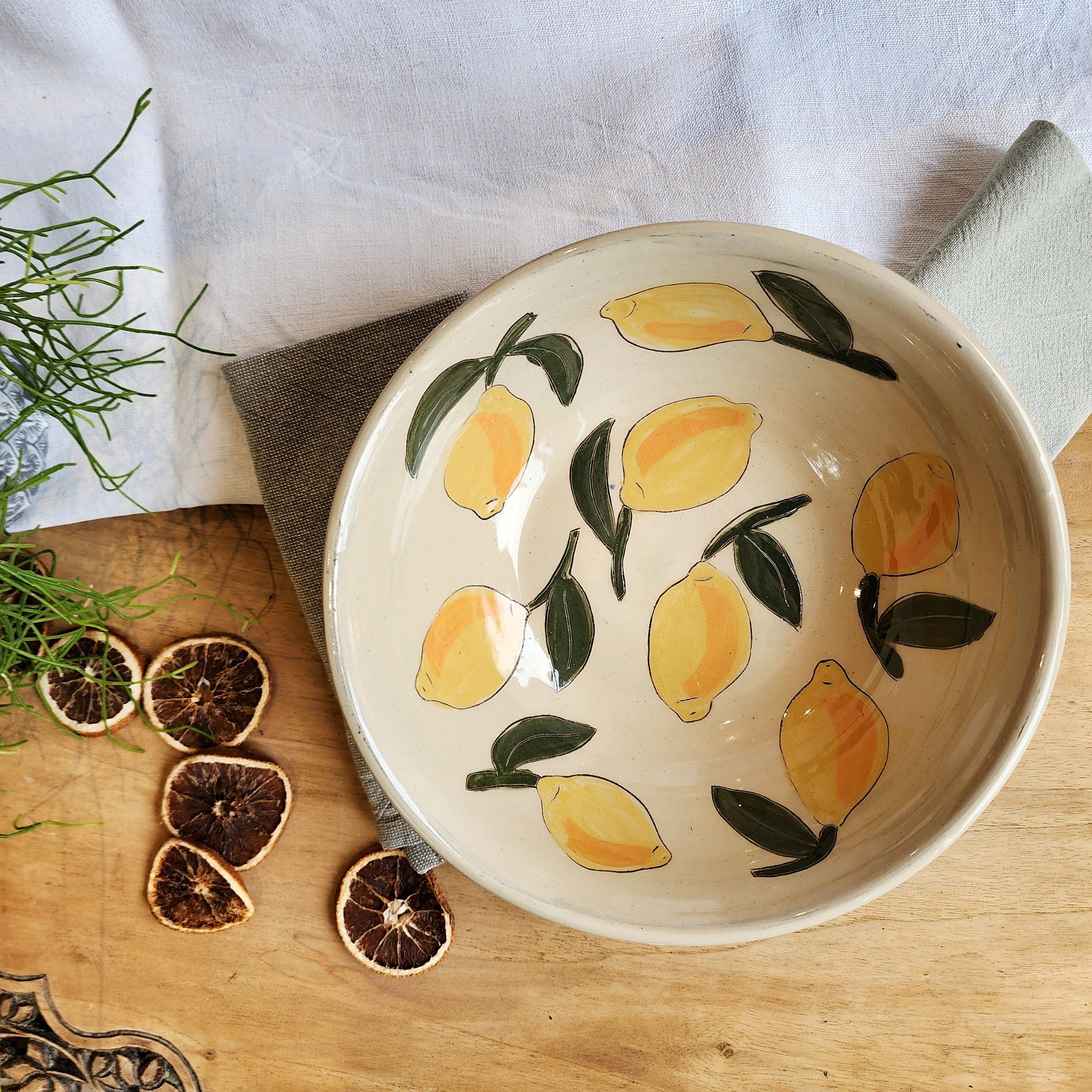 A ceramic bowl with a lemon and leaf pattern, placed on a wooden table with dried lemon slices and green plants nearby.