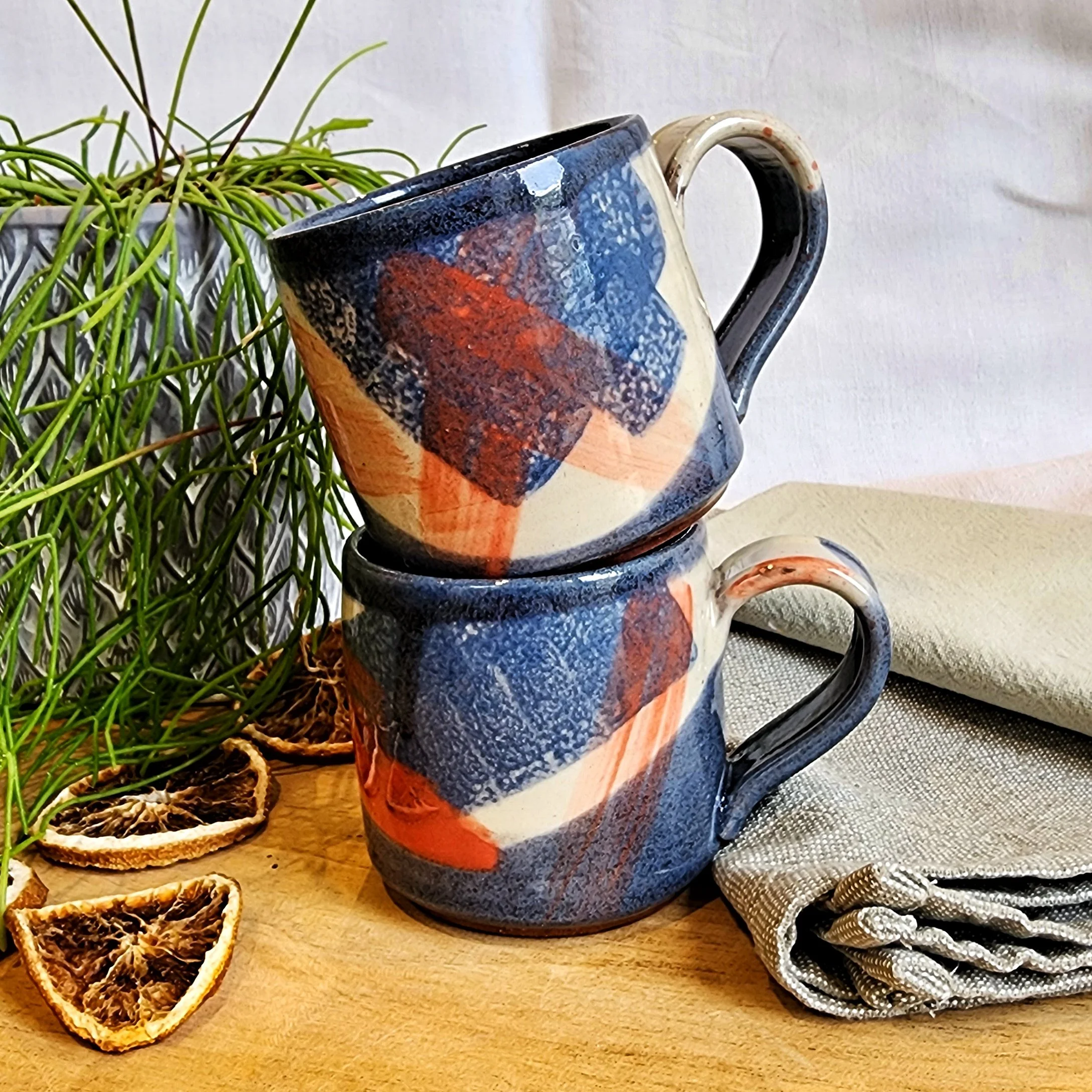 Two stacked ceramic mugs with abstract multicolored patterns, placed on a wooden surface surrounded by dried lemon slices, a green plant, a beige cloth, and a gray napkin.