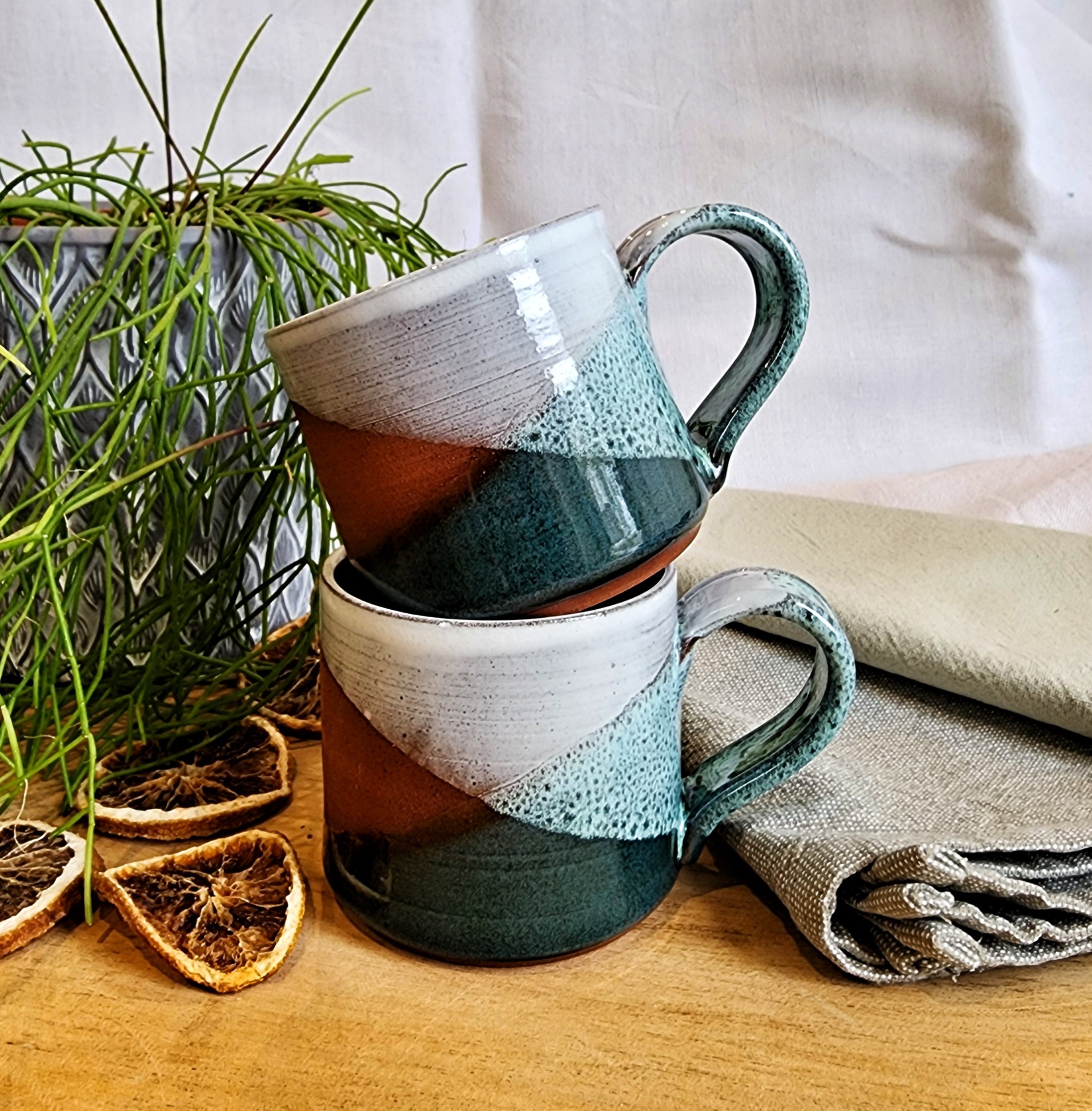 Stacked ceramic mugs with a color-block design, a potted green plant, dried citrus slices, and a folded cloth napkin on a wooden surface.