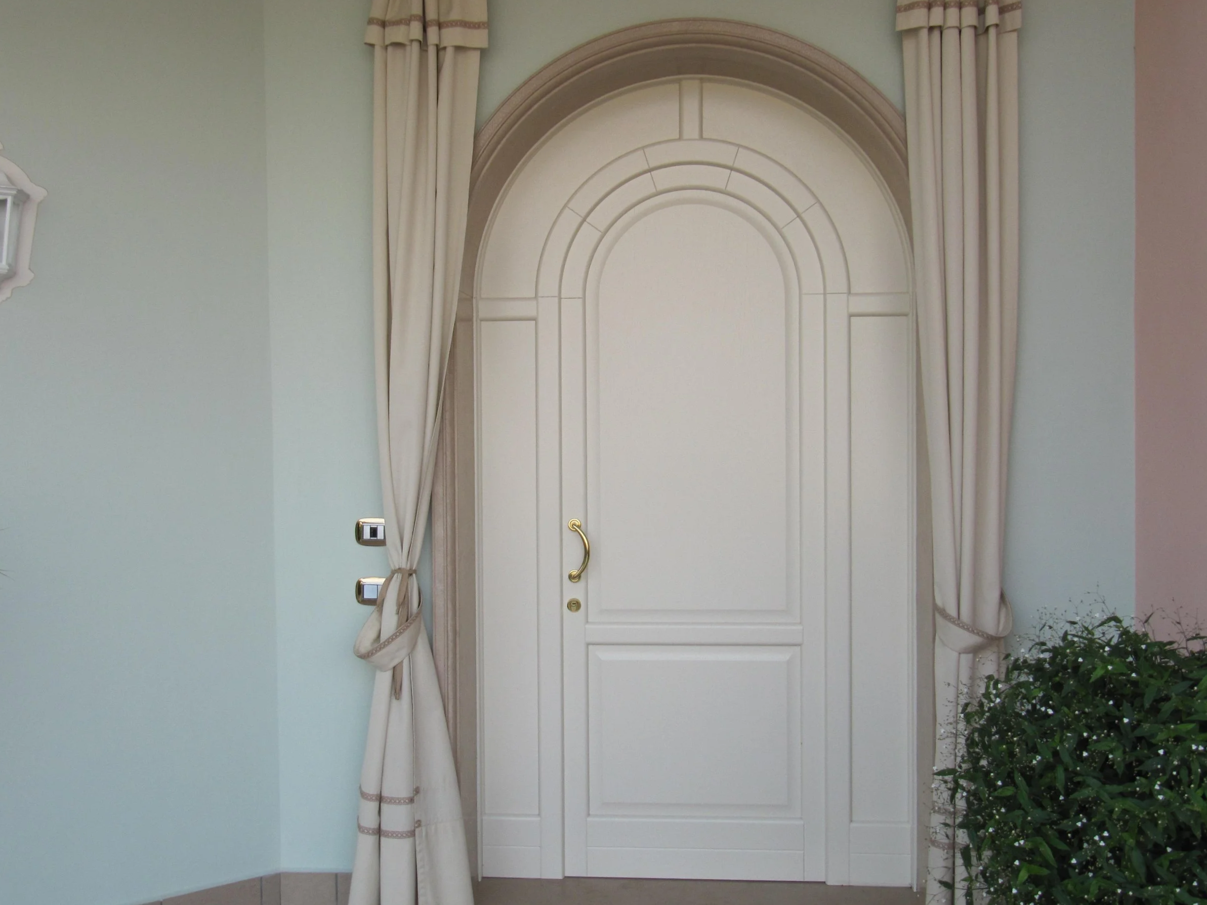 Porta bianca con arcata, cornici e maniglia dorata, circondata da tende beige con lacci, e con una pianta verde a destra.