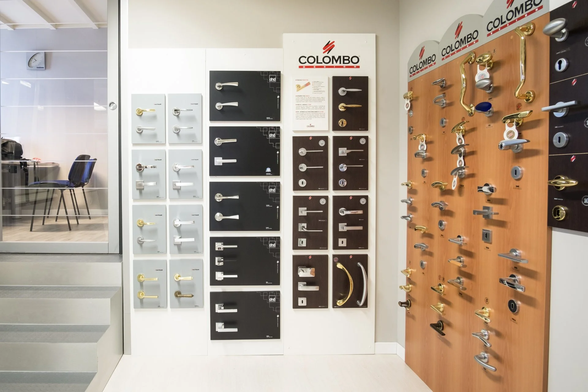 Mostra di maniglie e pomoli per porte in acciaio, ottone e altri materiali, esposti su pannelli di diversi colori e finish in un negozio di arredamento.