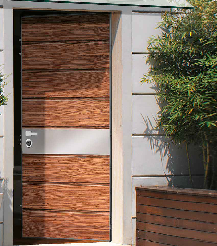 Porta moderna in legno con dettagli metallici e una serratura, circondata da muro in cemento e piante verdi.