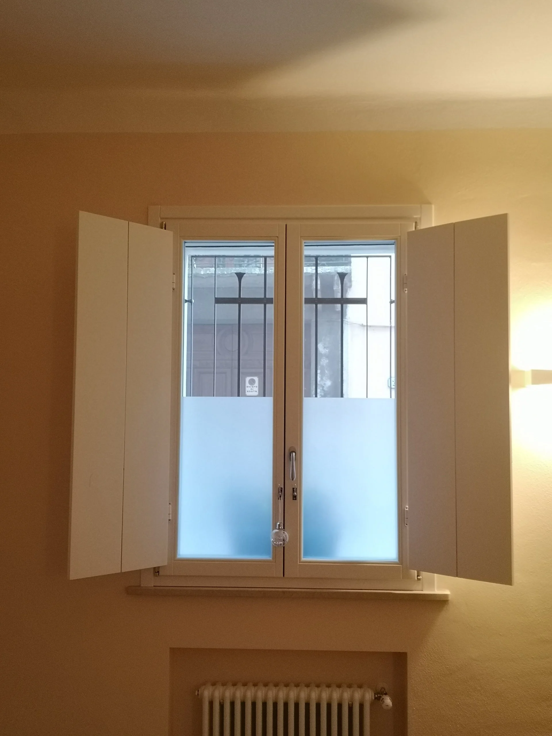 Finestra con ante chiuse su una parete gialla, con grate metalliche e vetri traslucidi, sotto una tapparella motorizzata