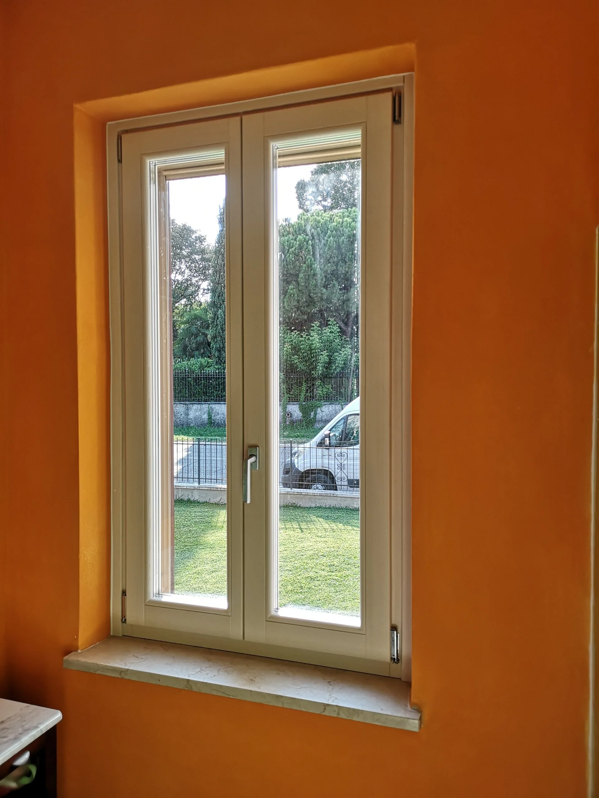 Finestra bianca a doppi vetri su una parete arancione, con vista su un giardino e una strada con una macchina bianca.