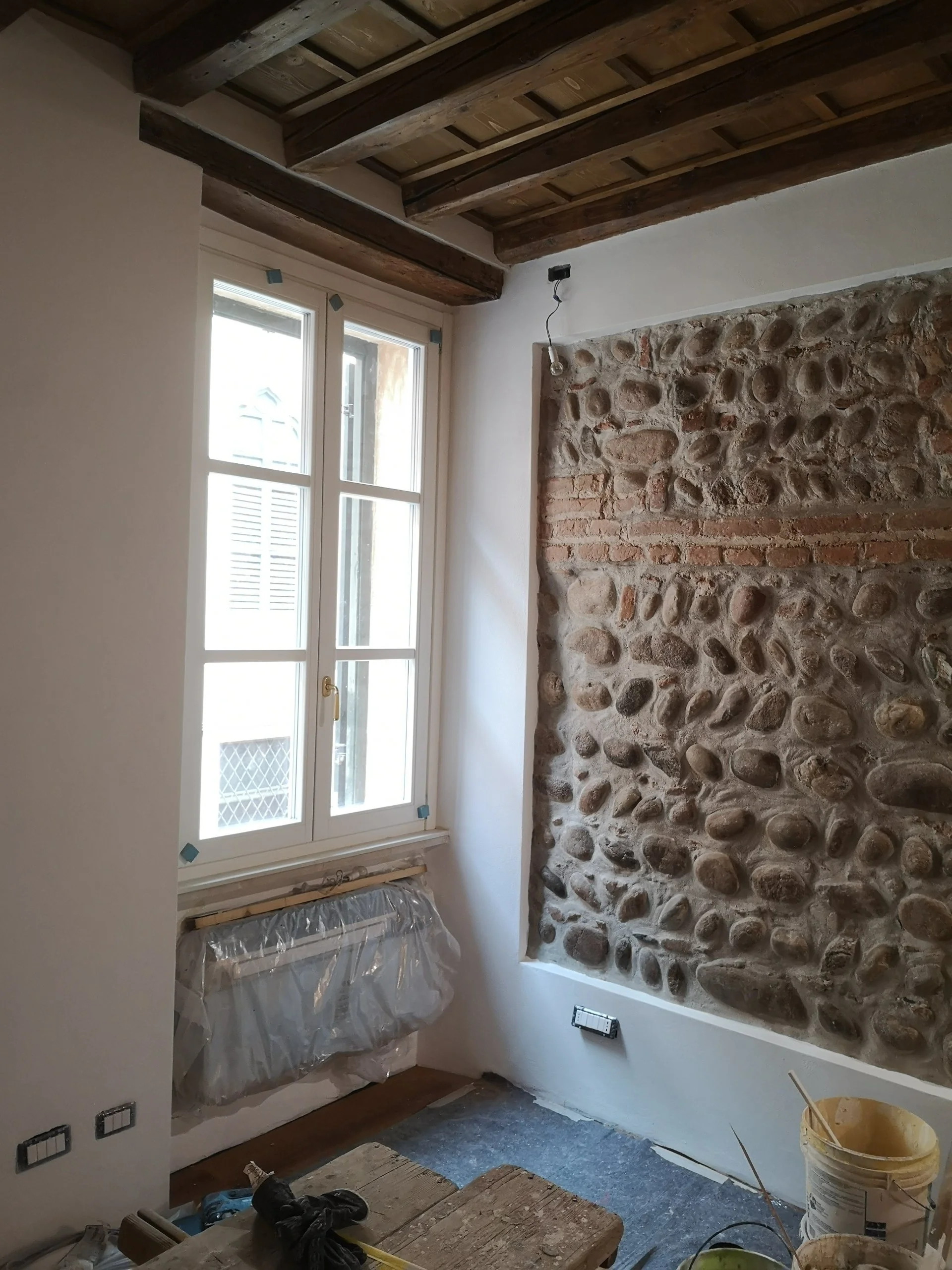 Interno di una stanza in fase di ristrutturazione, con parete in pietra e finestra, soffitto con travi in legno, e materiali di lavoro sparsi sul pavimento.