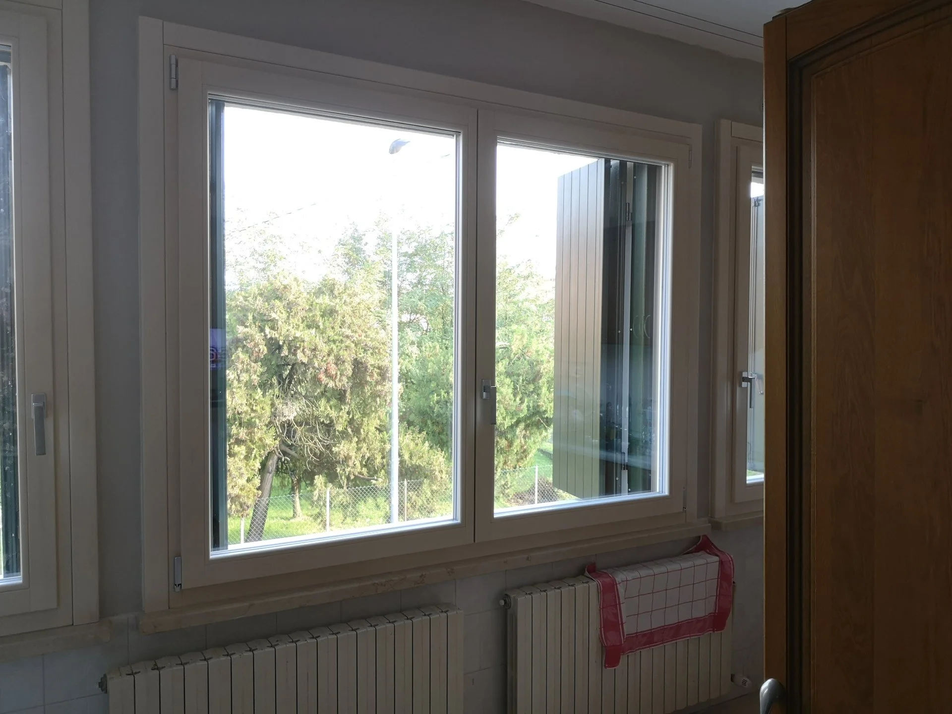 Finestra a doppia anta con vista su alberi e cielo, in una stanza con termosifoni sotto la finestra e muro bianco.