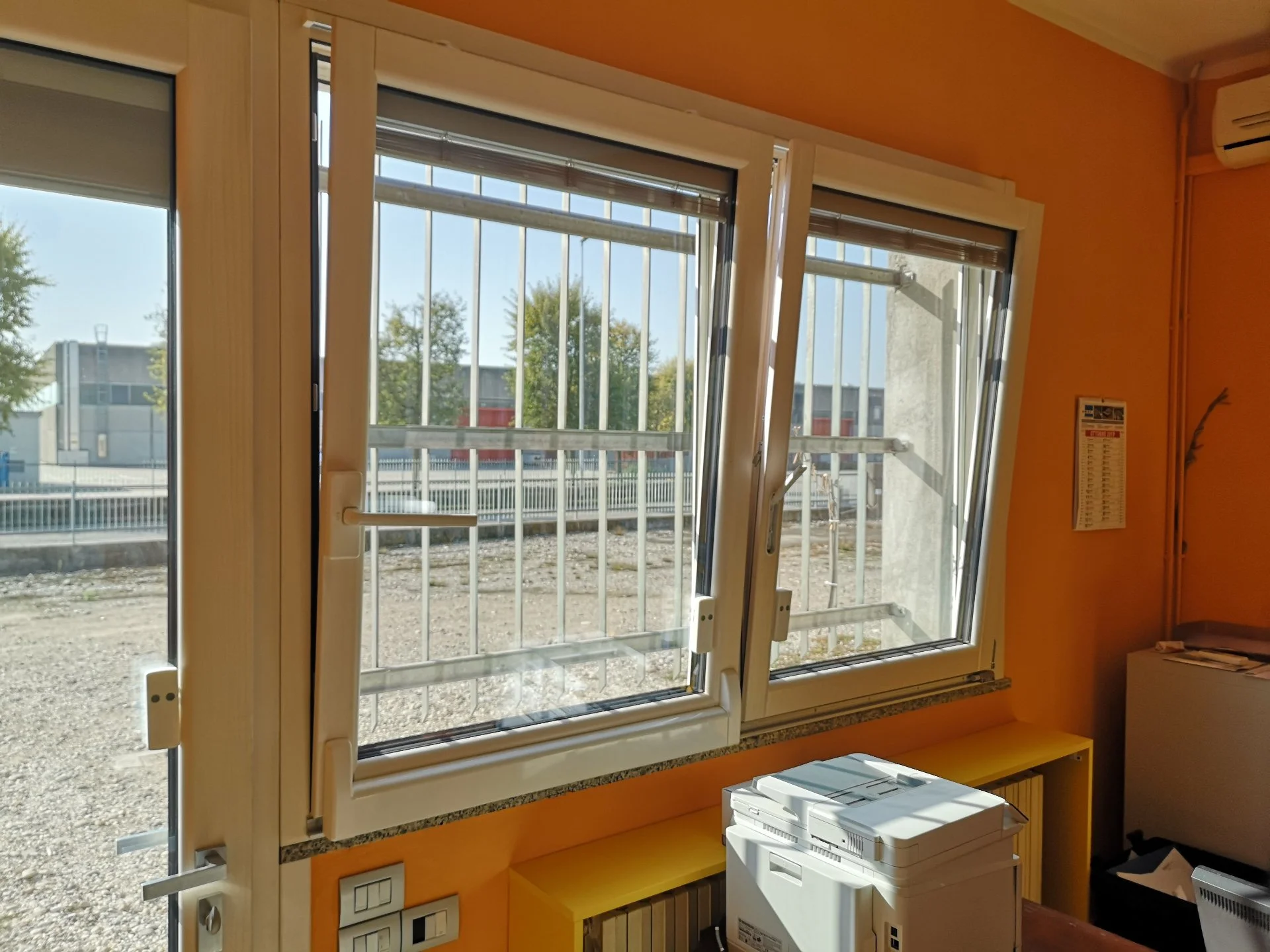 Finestra aperta con barre di sicurezza e vista su un'area esterna con edifici e alberi, all'interno di una stanza con pareti arancioni e arredo.