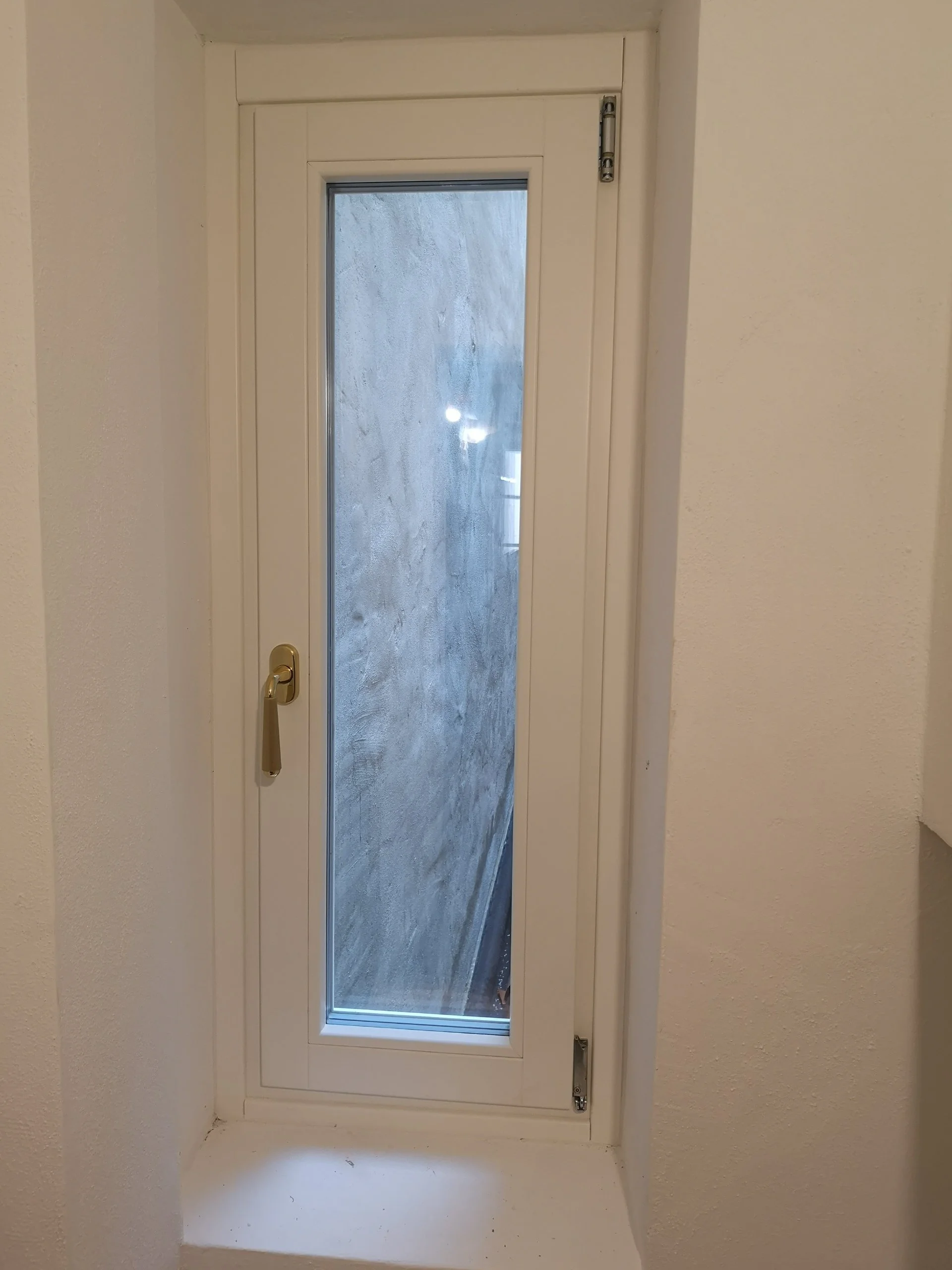 Finestra bianca con vetro trasparente, incorniciata da un telaio bianco, con maniglia d'oro e due cerniere, situata in una parete bianca, con una superficie di appoggio sotto.