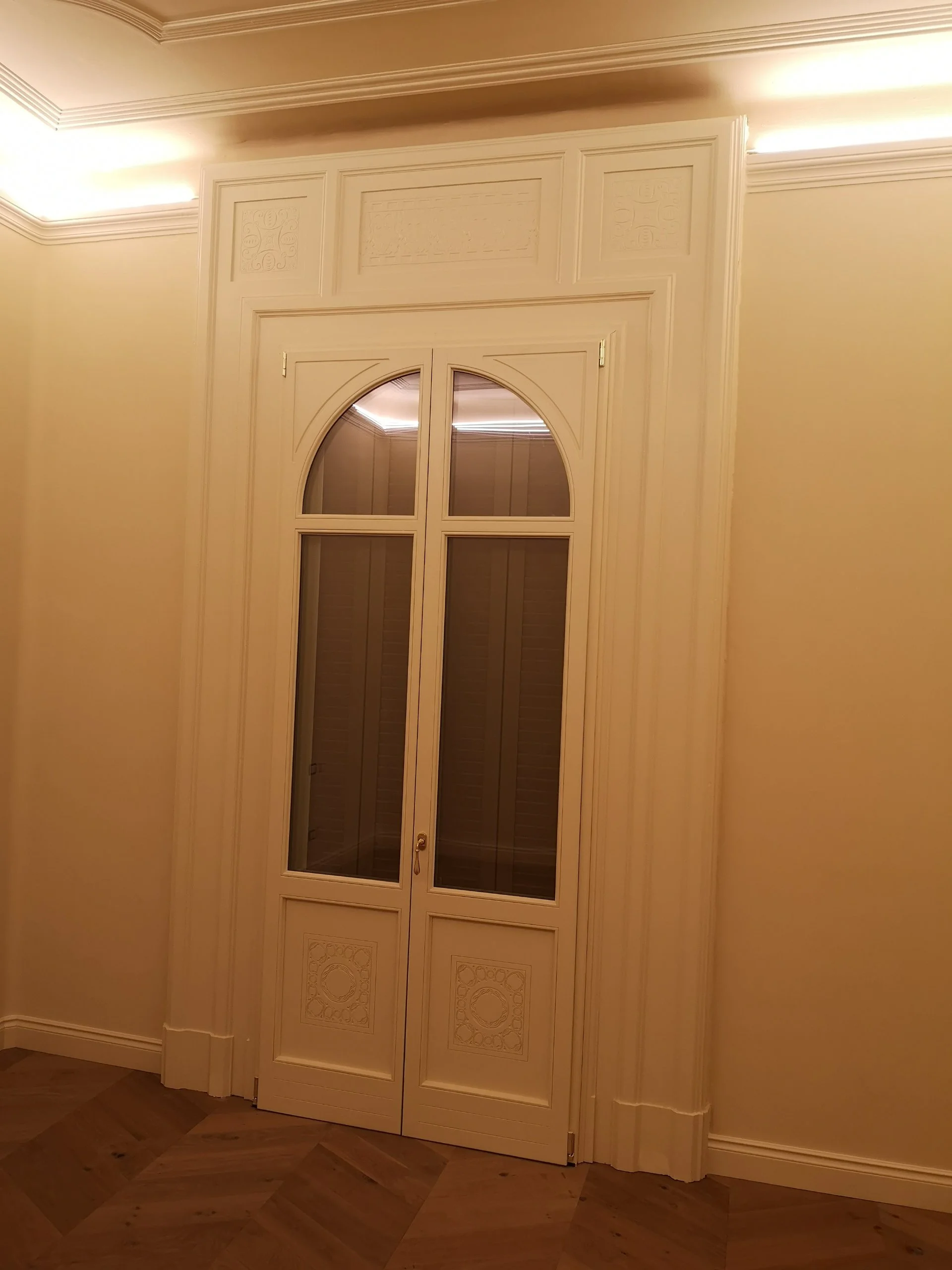 Una porta decorativa in stile classico con vetri e dettagli intagliati, incorniciata da ornamenti e pannelli di parete chiari, con un pavimento in legno a motivo geometrico.