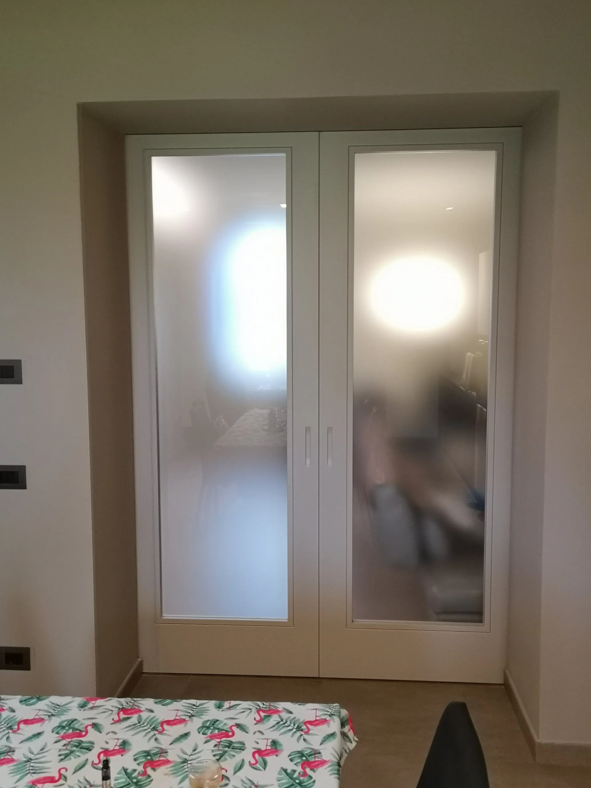 Vetrate in vetro smerigliato di una porta bianca, parzialmente visibile un tavolo con tovaglia decorata con fenicotteri rosa e foglie verdi, e alcune sedie in un ambiente domestico.