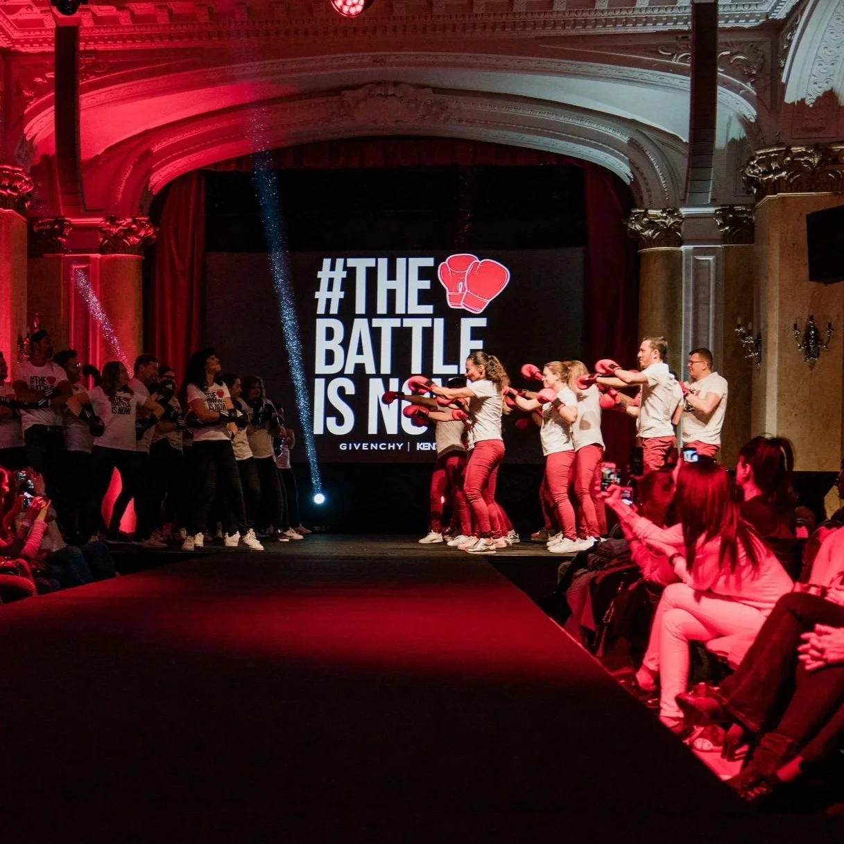Une scène de défilé de mode ou d'événement organisé avec un groupe de personnes en tenue rouge et blanche, certaines en train de faire une démonstration ou une performance sur scène. À l'arrière-plan, un grand écran affiche le hashtag #TheBattleIsOn avec des gants de boxe rouges et la mention Givenchy, Ken.