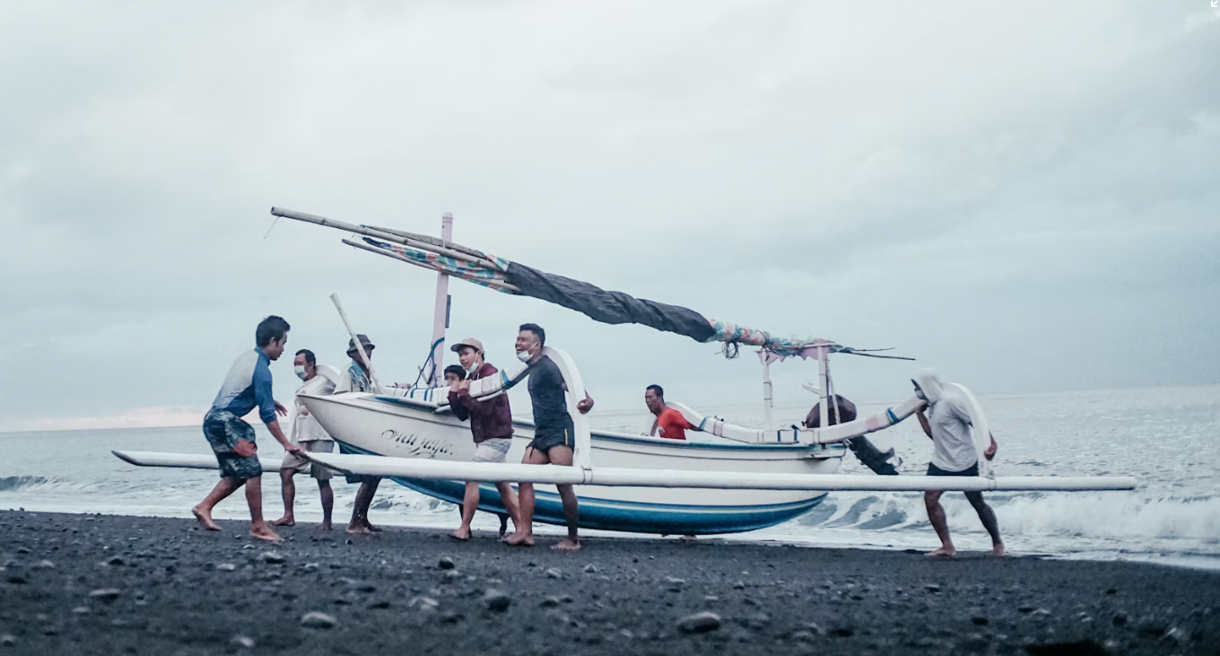 Groupe de personnes portant un bateau de pêche sur une plage, mer calme en arrière-plan, ciel nuageux.