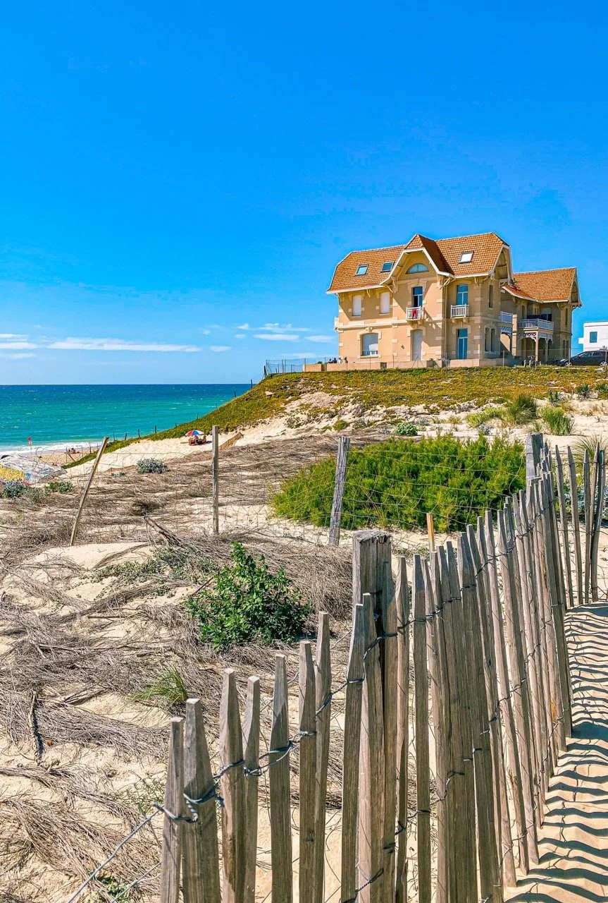 Maison de style traditionnel avec toit en tuiles rouges sur la dune côtière de Biscarrosse. La vue comprend la plage, l’océan Atlantique et une clôture en bois rustique au premier plan.
