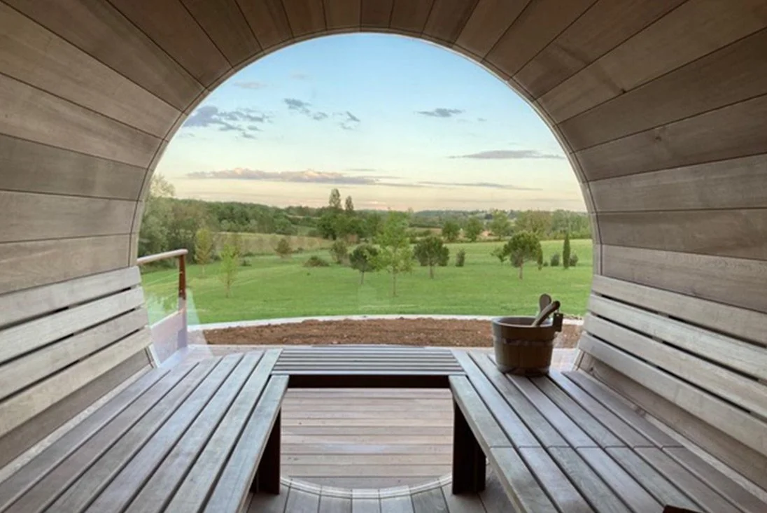 Vue d'un sauna en bois avec un banc en bois à l'intérieur, offrant une vue sur un paysage verdoyant avec des arbres et un ciel partiellement nuageux.